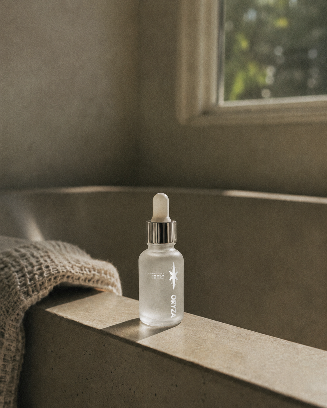 A frosted glass dropper bottle labeled 'ORYZA' sits on a beige surface near a window with natural light. A textured blanket is draped over the edge of the surface. The scene has a cozy, minimalist aesthetic.
