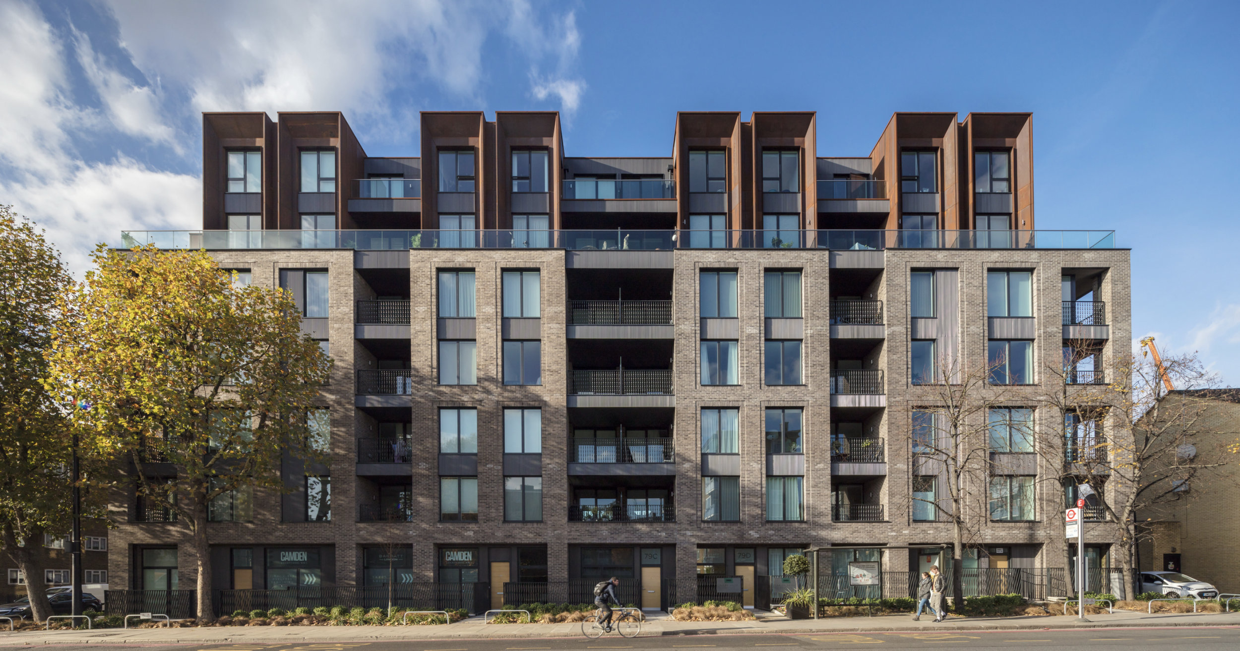 Modern multi-story apartment building with brick and glass facade, trees in front, and a blue sky with clouds.