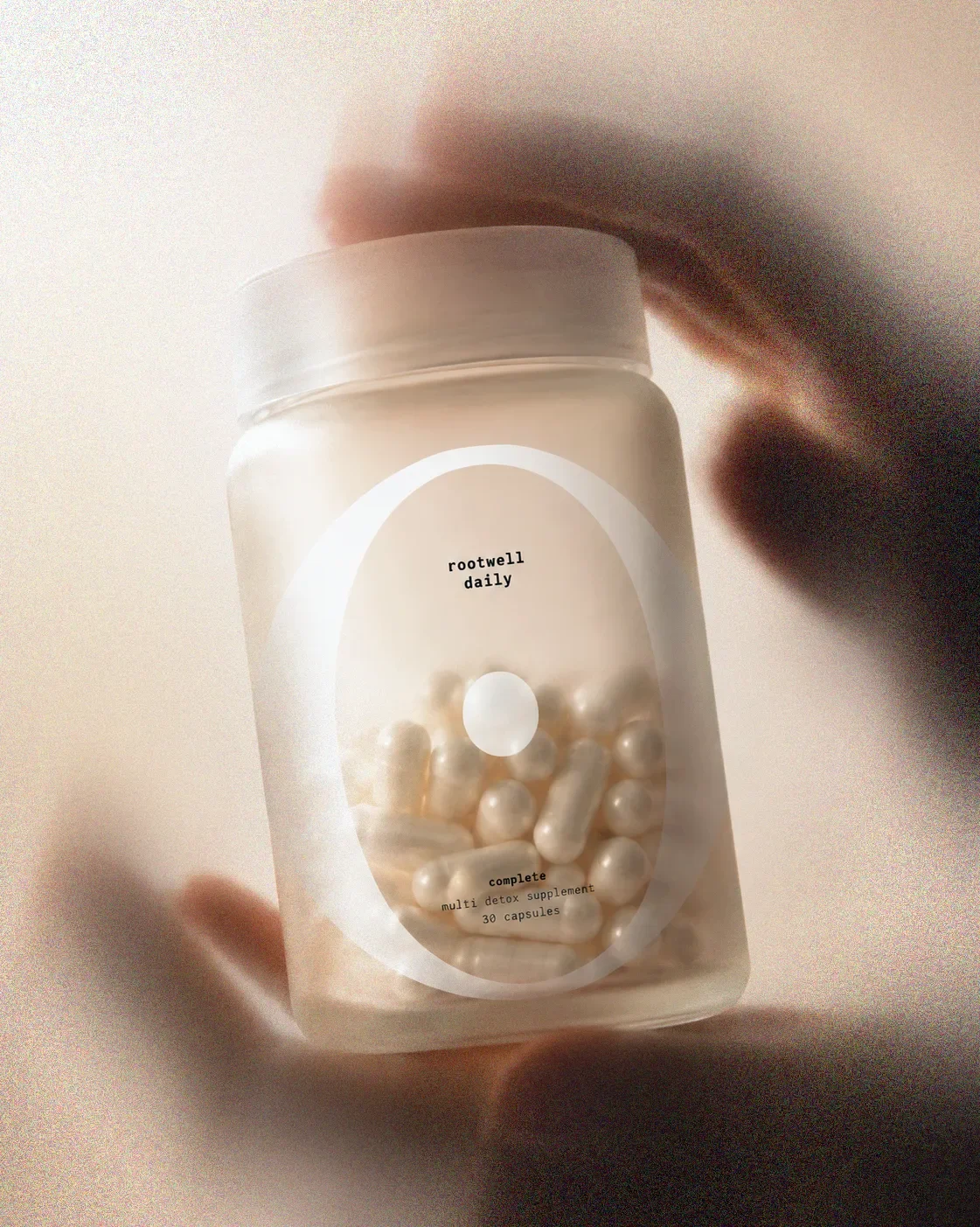 A hand holding a clear plastic container of Rootwell Daily multivitamin capsules, with a white label featuring minimalist design and small black text.