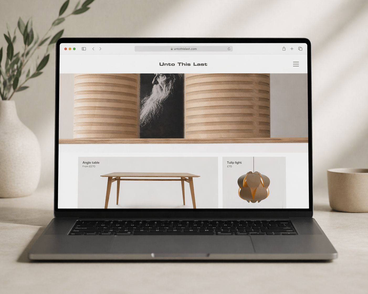 Laptop on a desk displaying a webpage for furniture and lighting, with a plant and a cup nearby.