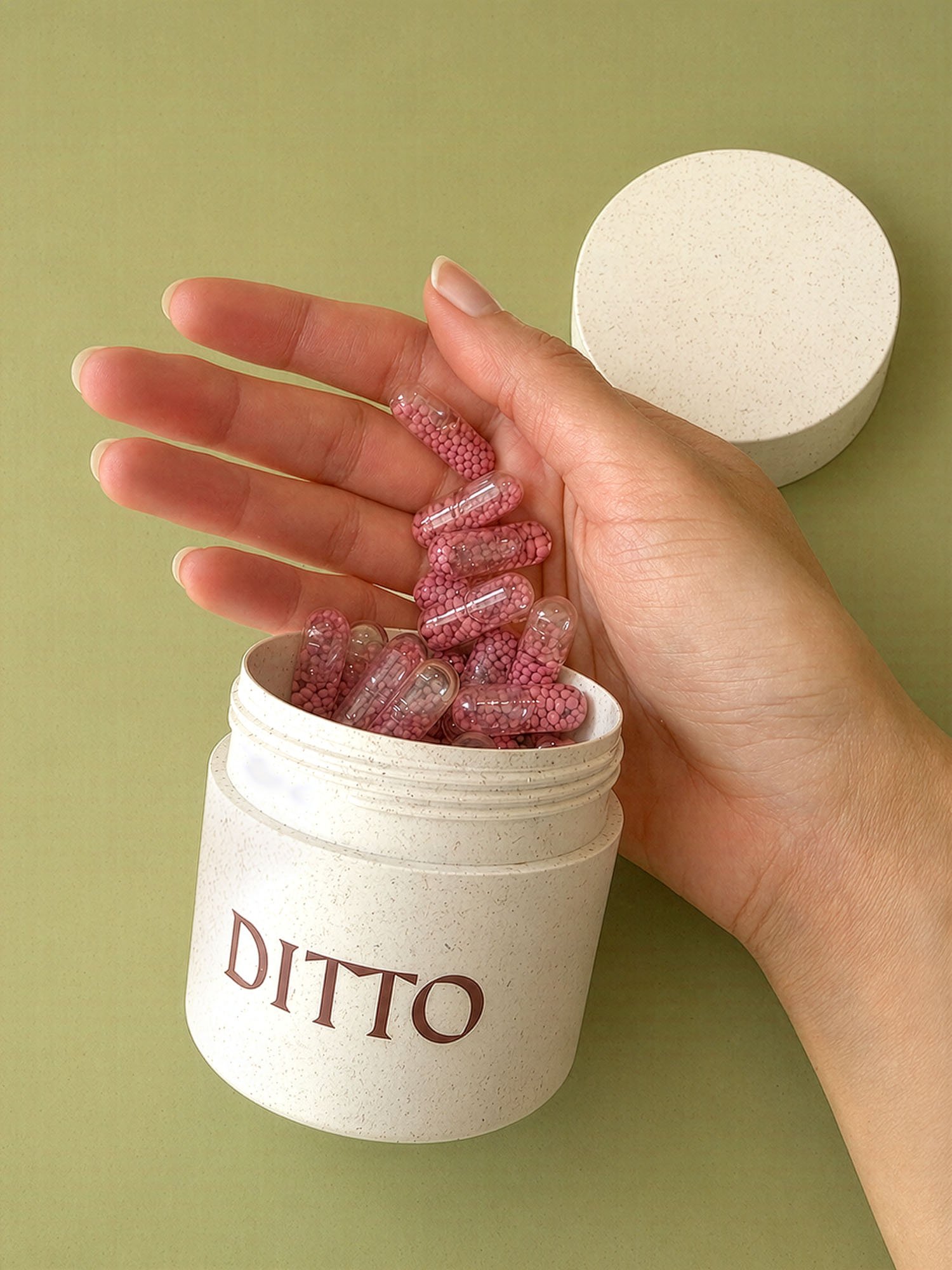 A person's hand with gel capsules on fingers, and a container with similar capsules, next to a white jar with the word partially visible on the label, set against a green background.