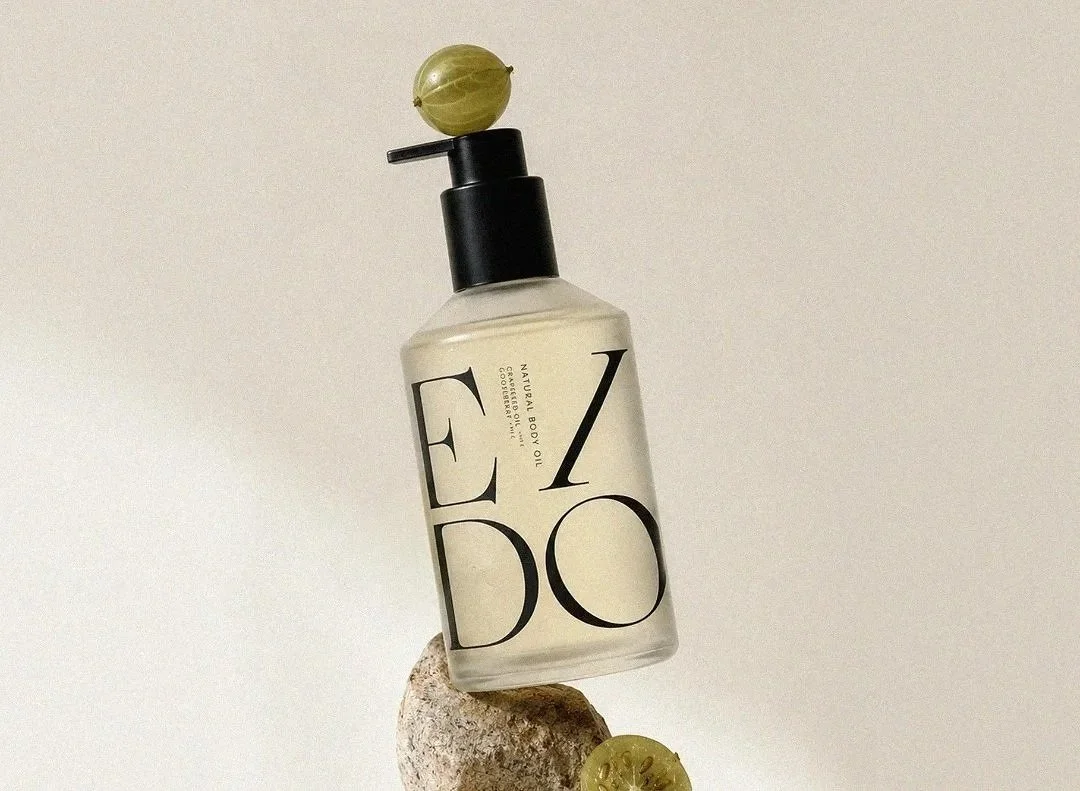 A bottle of body oil placed on a small rock, with slices of green gooseberry around it and a whole gooseberry balanced on top of the pump, all against a plain neutral background.