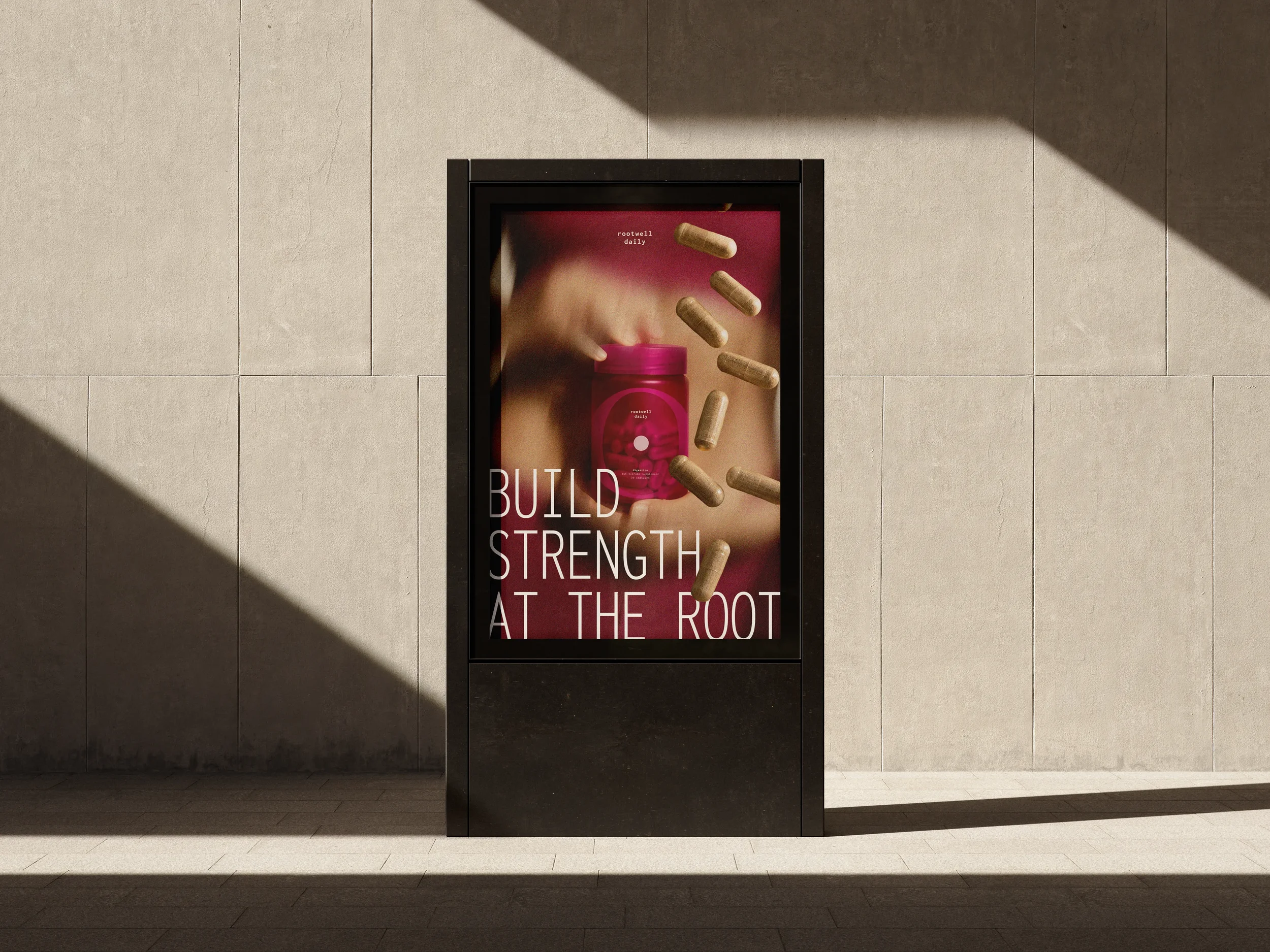 Digital advertisement on a wall with a pink container and capsules falling around it, with text about building strength from the rootwell daily brand.