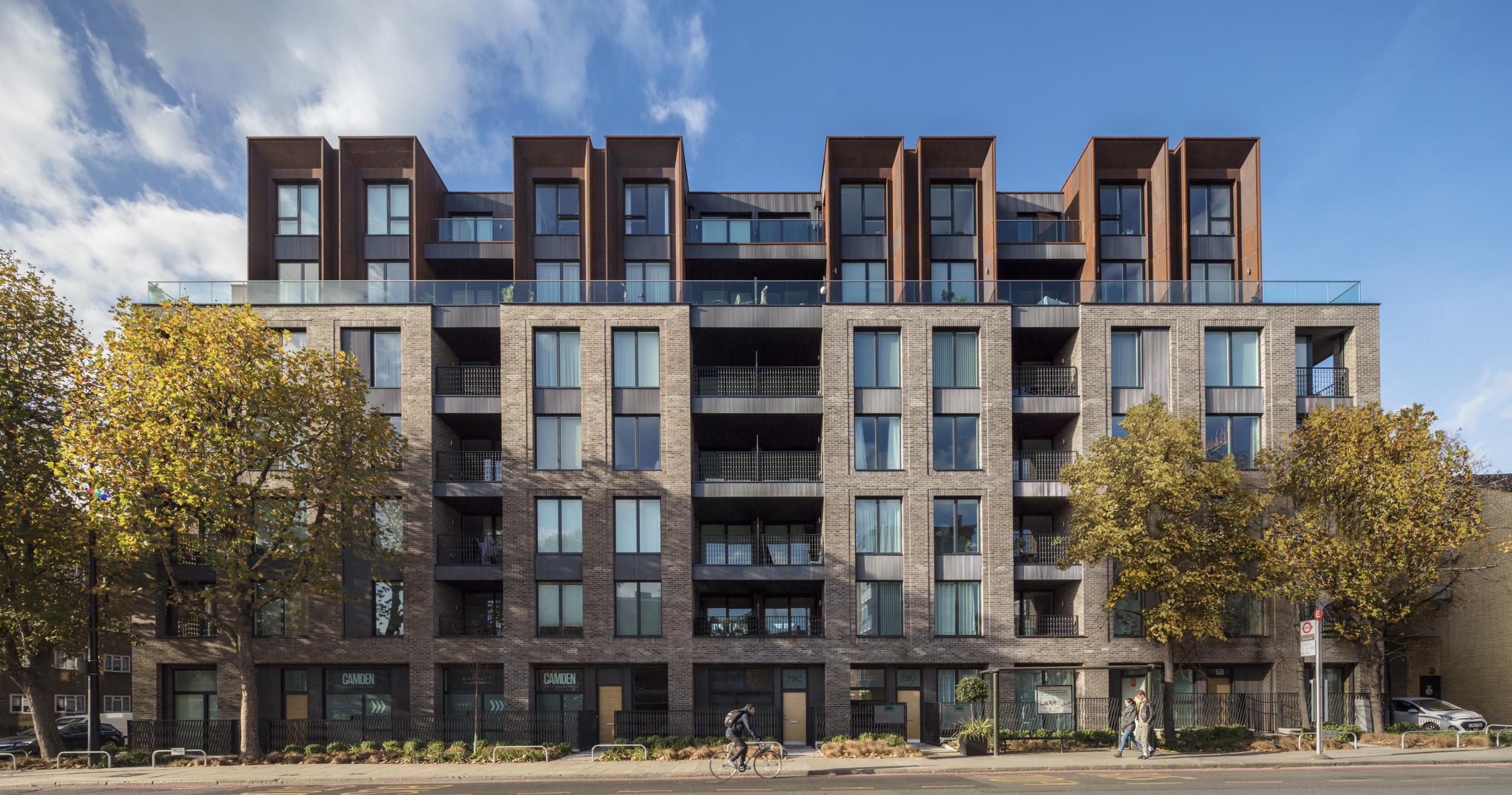 A multi-story modern apartment building with brick and metal exterior, glass balconies, and large windows, with trees and people walking or cycling in front under a partly cloudy sky.