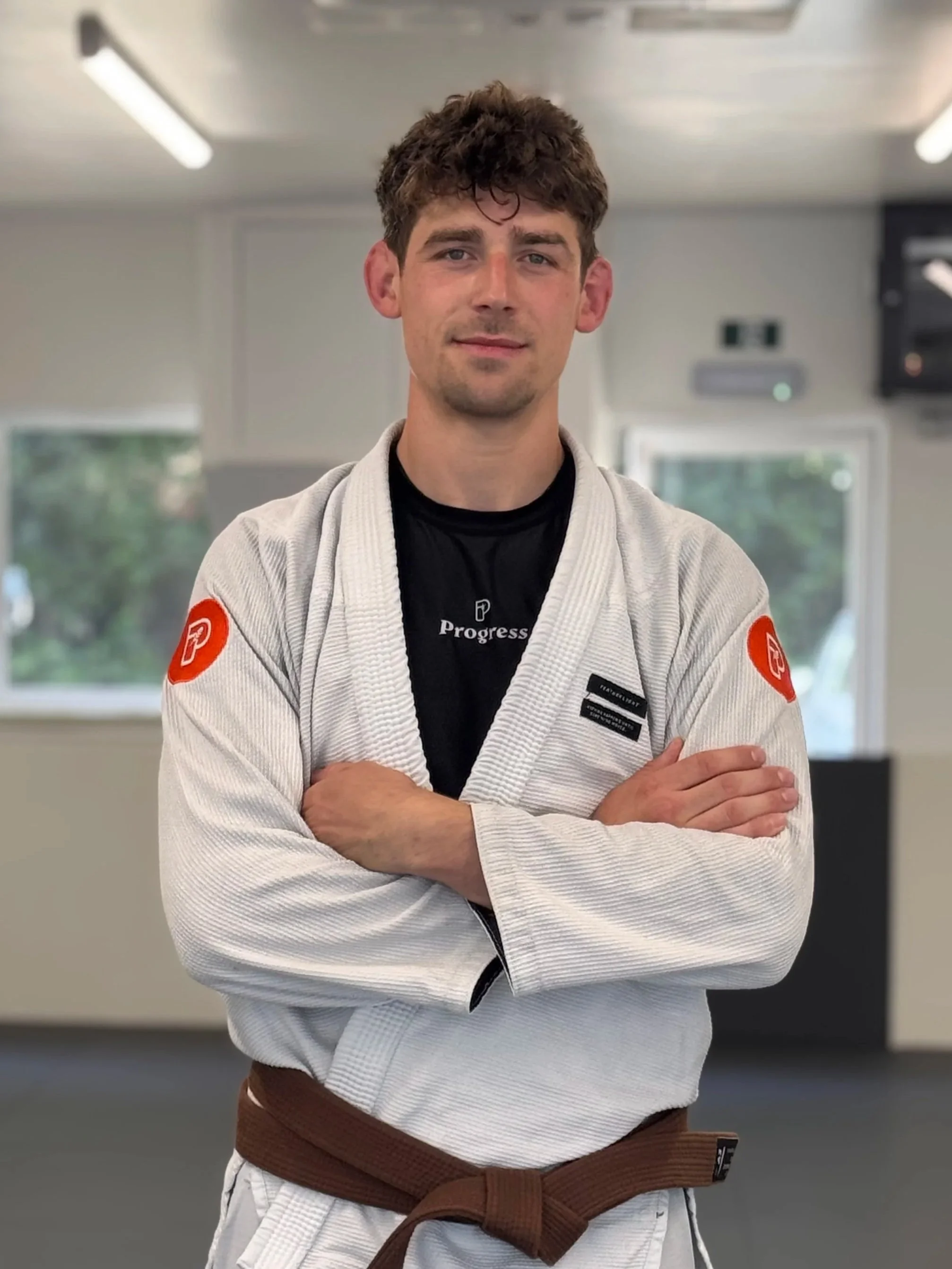 A man in a black martial arts gi with a brown belt, standing with hands on hips in a training room.