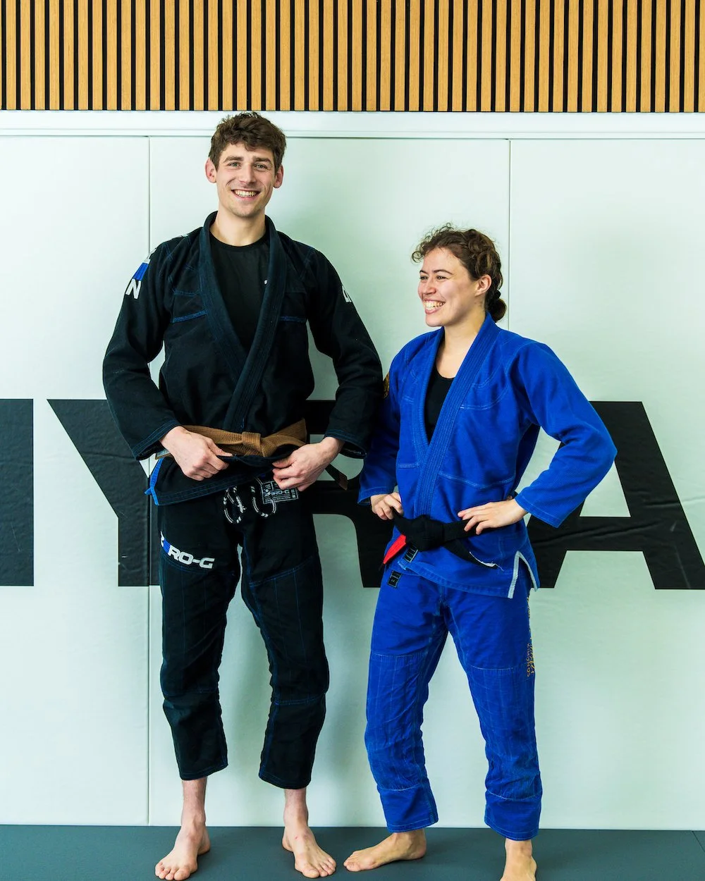 Two people in Brazilian Jiu-Jitsu gi uniforms standing on a mat inside a training facility, smiling and posing for a photo.