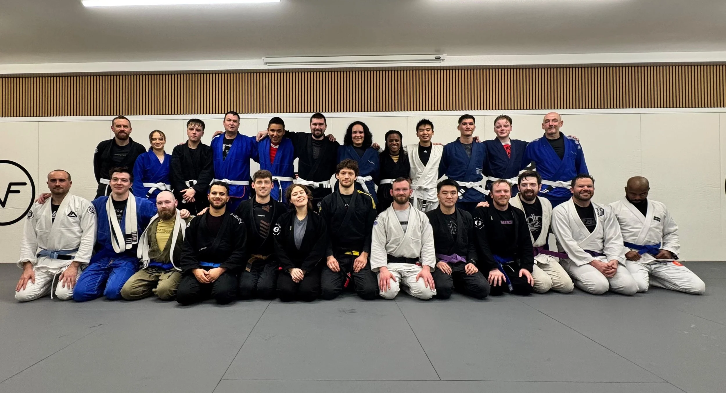 Group of people in Brazilian Jiu-Jitsu gis and belts posing in a martial arts training facility.