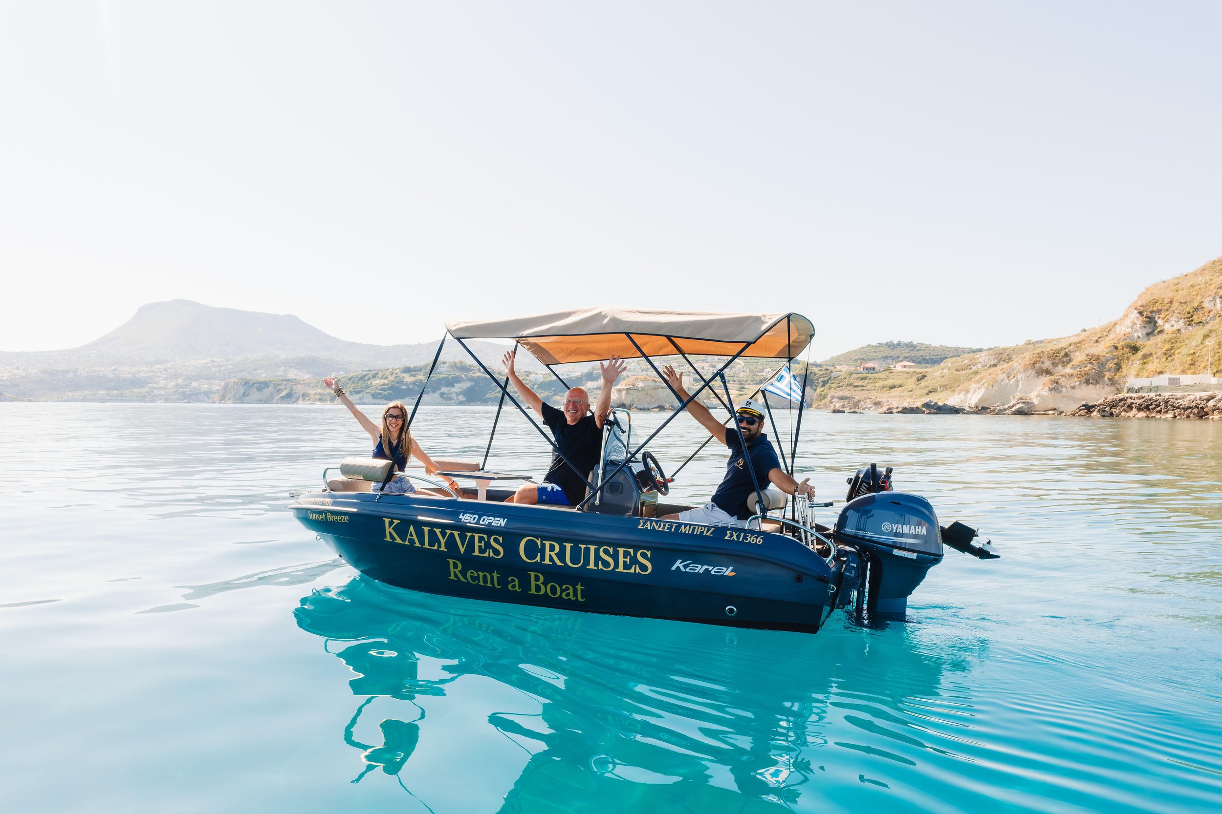 Three people on a blue motorboat on calm water with cliffs and hills in the background. They are smiling and raising their arms, enjoying a sunny day. The boat has signage advertising Kalyves Cruises and boat rental.