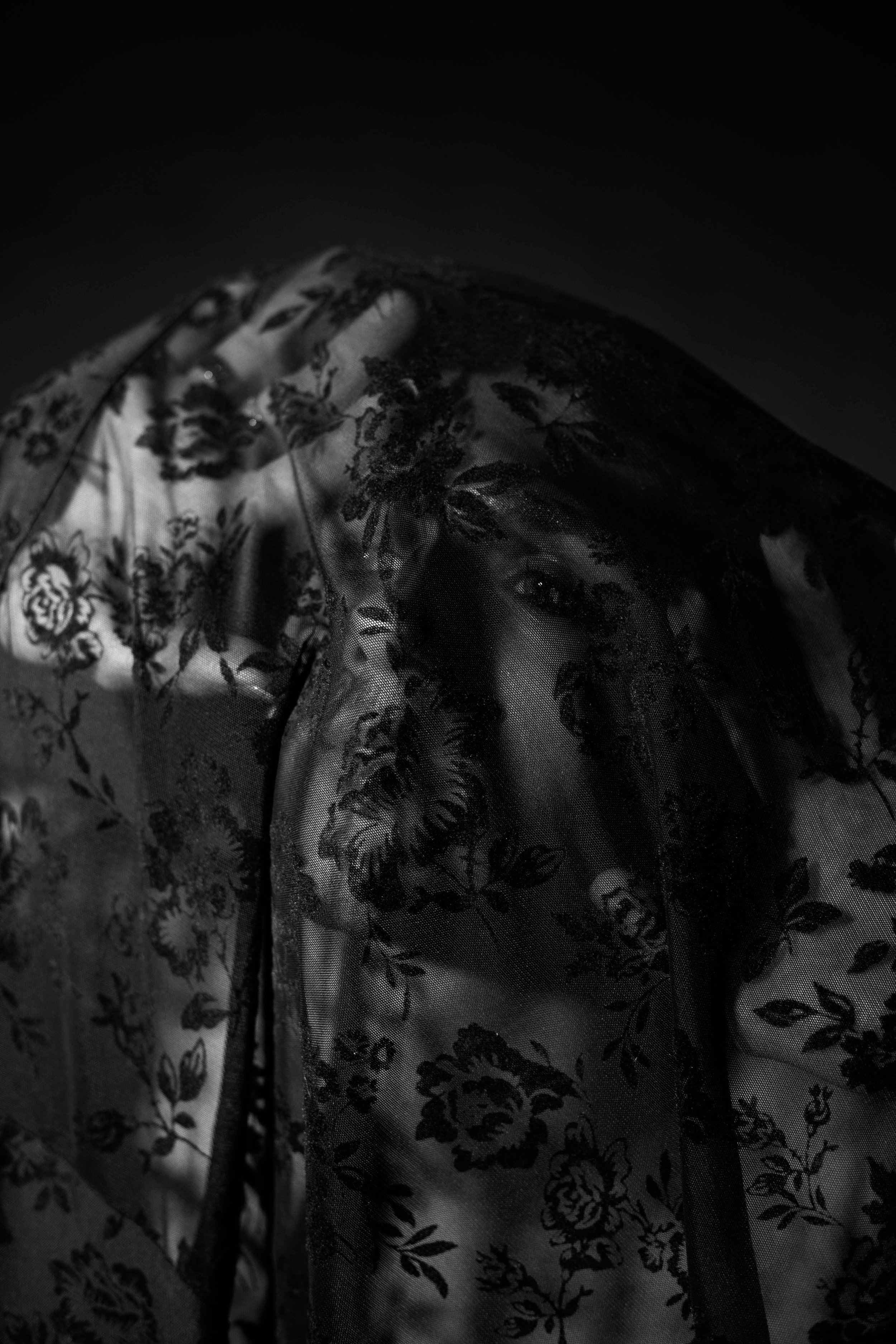 A black and white photo of a man partially hidden behind a sheer floral patterned curtain or fabric. Only part of her face is visible through the fabric.