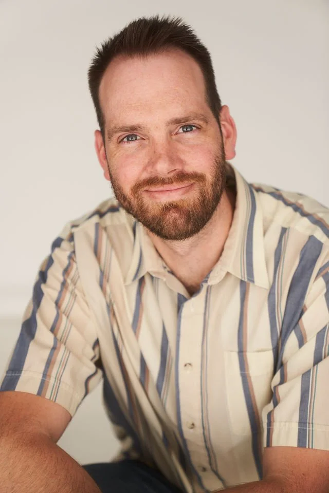 Un homme avec une barbe et des cheveux courts, portant une chemise à rayures, souriant devant un fond blanc.