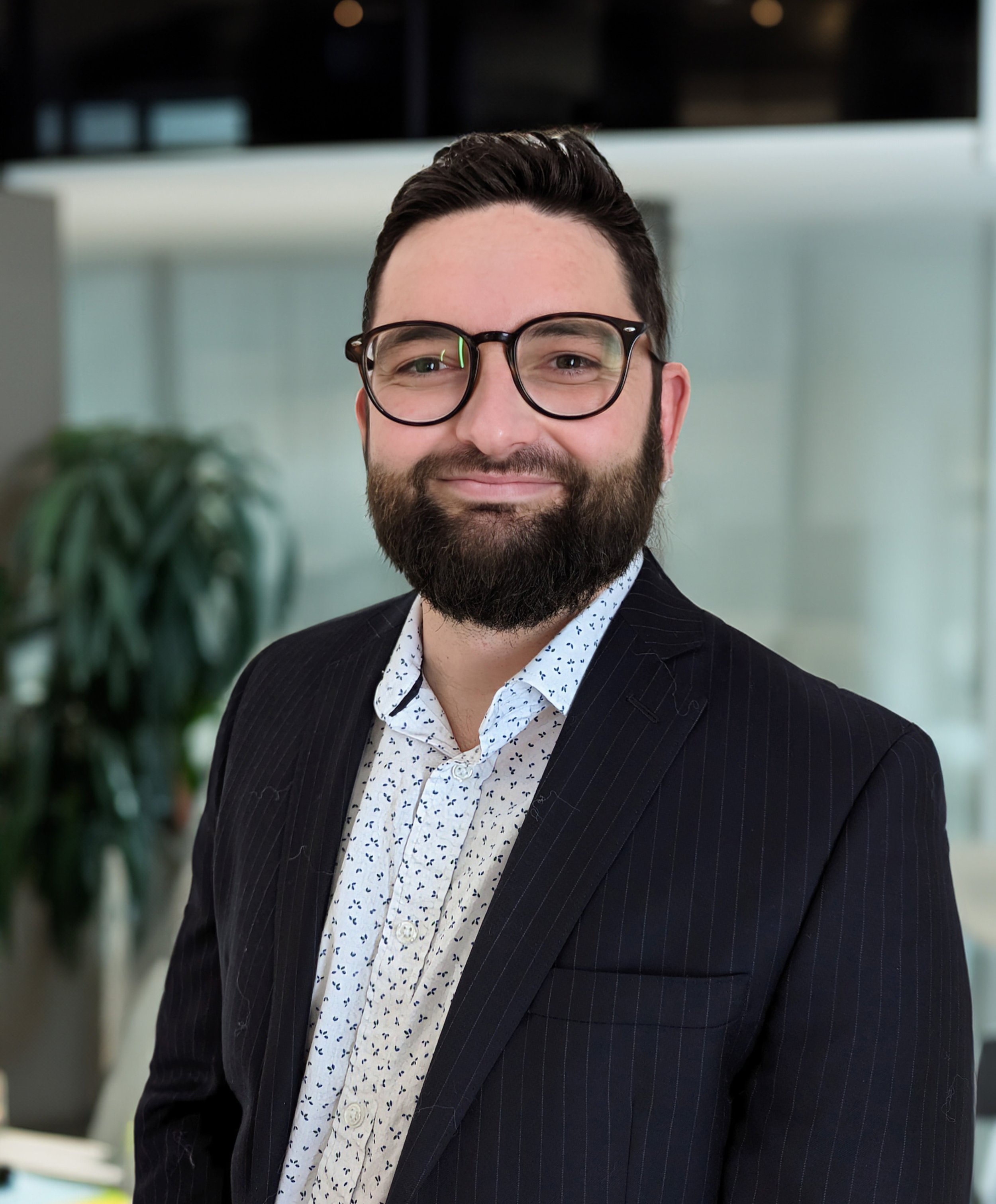 Portrait d'un homme souriant, portant des lunettes, une chemise blanche à motifs, et une veste noire à rayures, dans un environnement professionnel moderne.