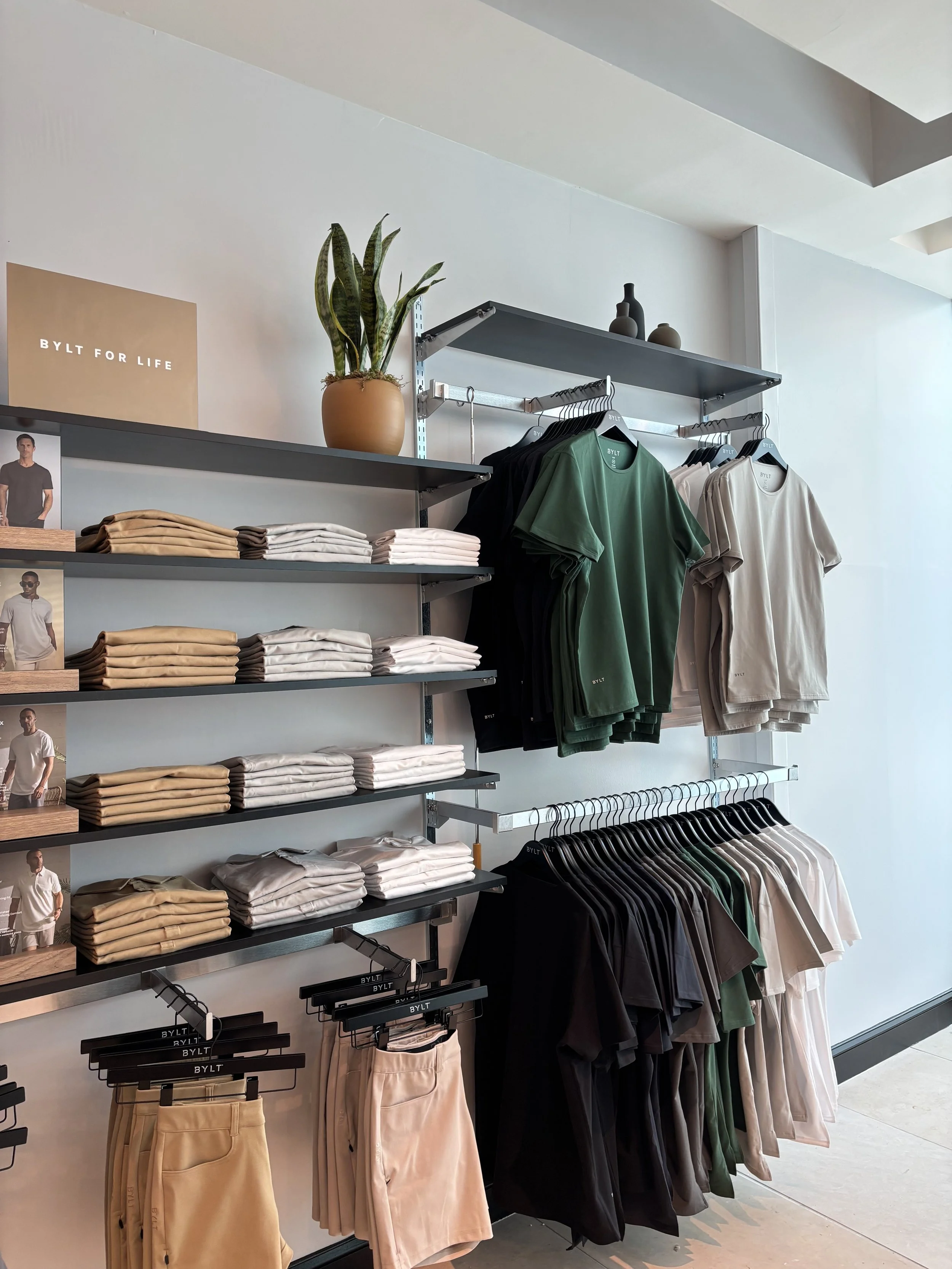 Photo of a display in a clothing store with shirts and shorts and a houseplant on the top shelf.