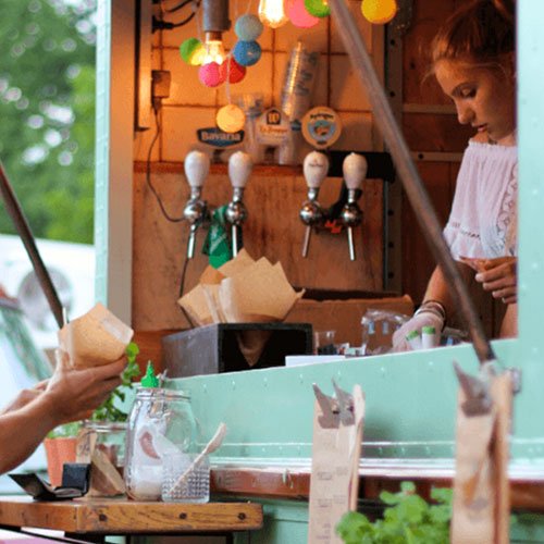 A smiling woman in a blue and white dress and hat sitting next to a man in a striped blazer at an outdoor event, holding drinks and sitting at a table.