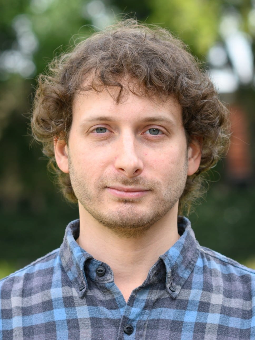 Close-up of a young man with curly light brown hair and a beard, looking at the camera in an indoor setting.