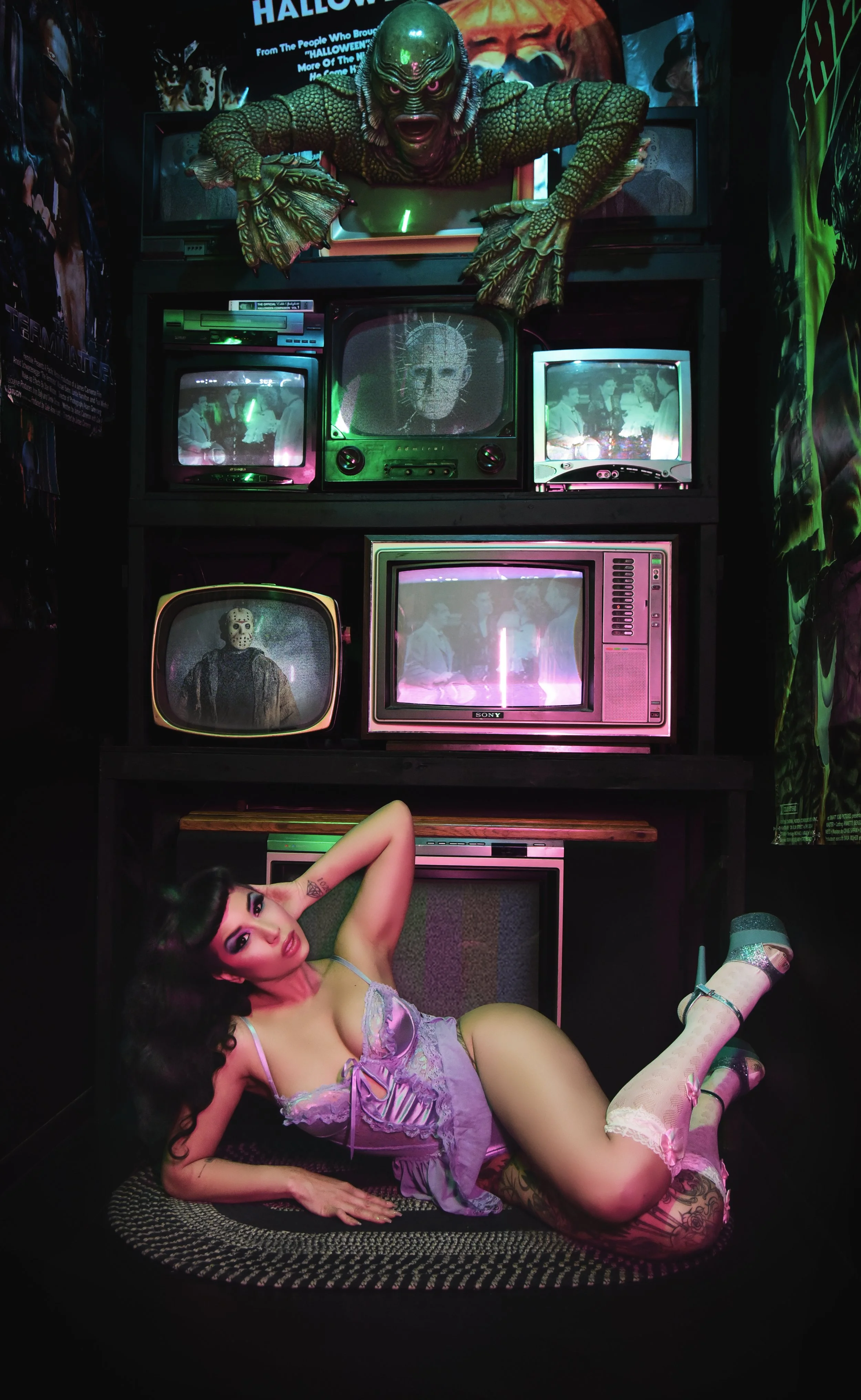 Lou Lou la Duchesse de Rière is an Afro Indigenous performer, shown in a purple costume, reclining in front of a wall of old TVs showing horror movies. 