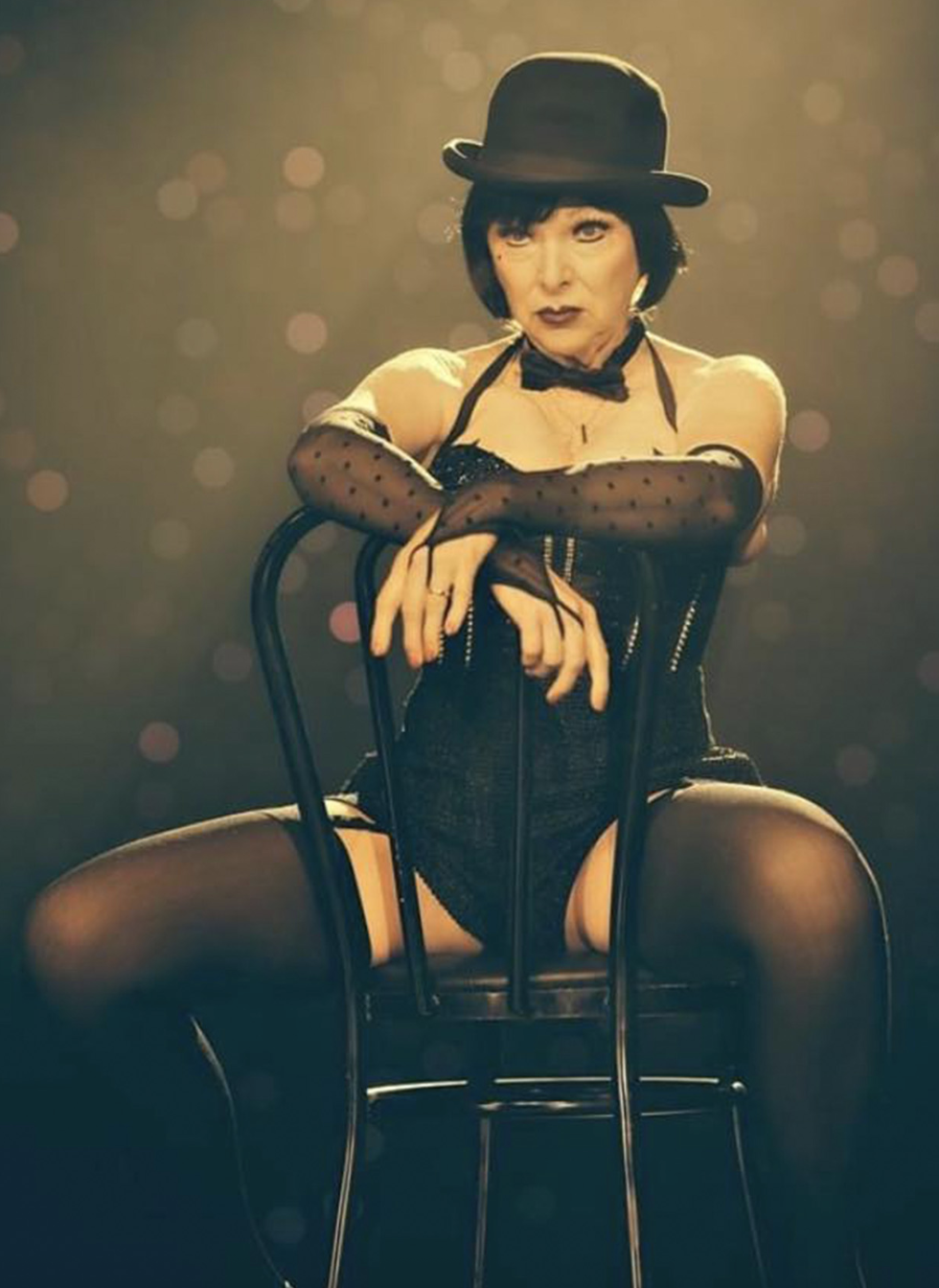 Lovey Goldmine straddles a chair and is wearing a Cabaret inspired costume