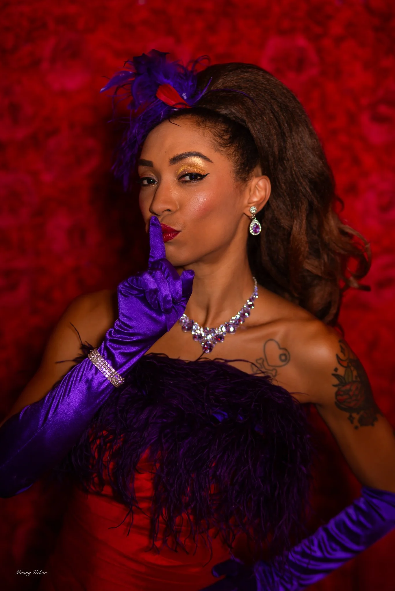 Jeez Loueez, a petite black performer wearing a gown trimmed in ostrich feathers. She holds one finger over her mouth in a silence gesture.