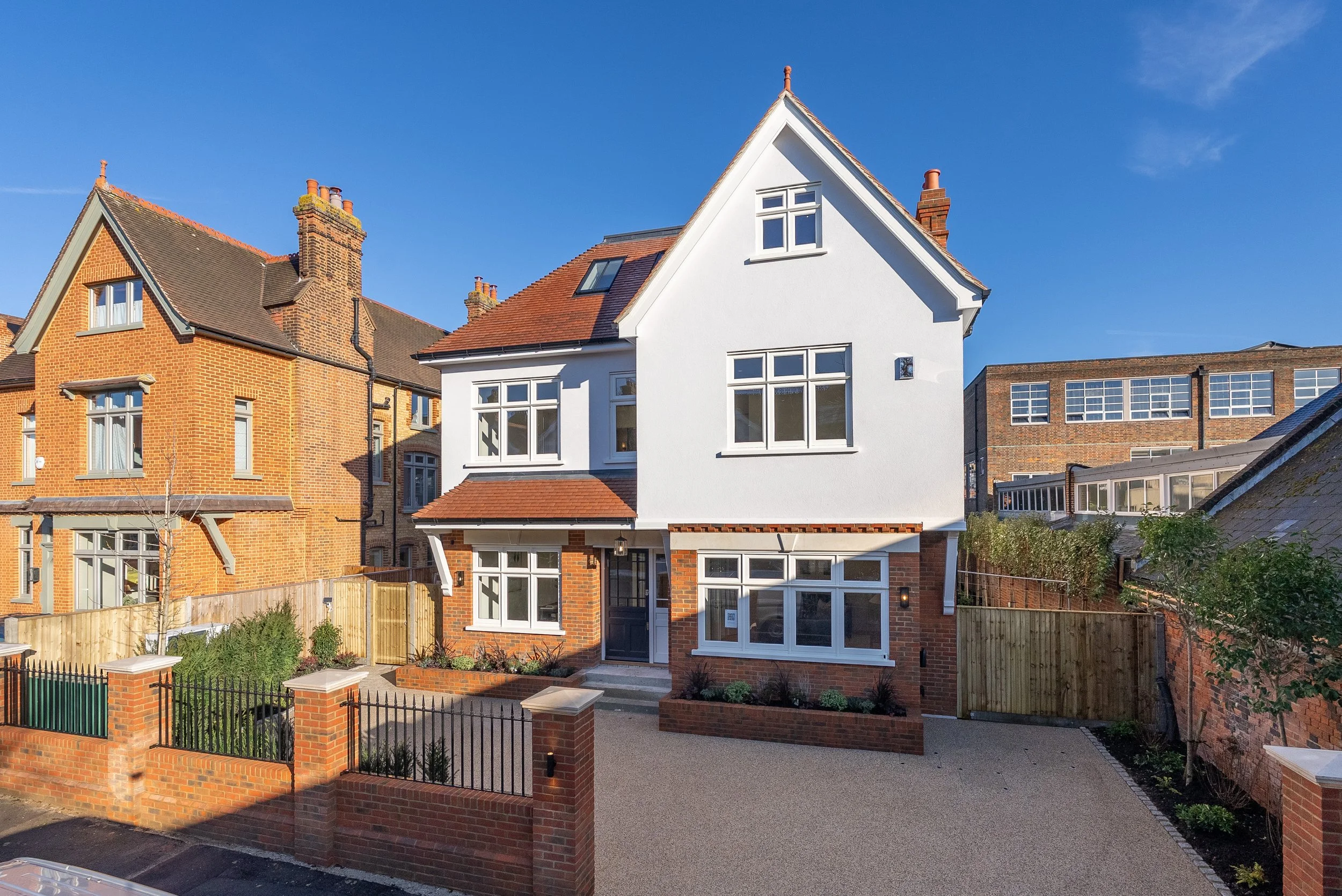 A white, three-story + Lower Ground Luxurious house situated in a Wimbledon Village in close proximity to Kings College, a fenced front yard, and a clear blue sky.