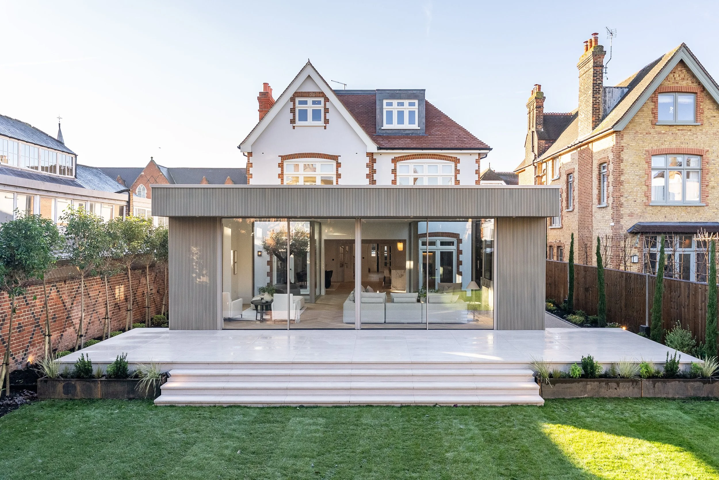 Modern house with large glass windows and a patio, surrounded by a green lawn and wooden fences, in a neighborhood with traditional brick houses.
