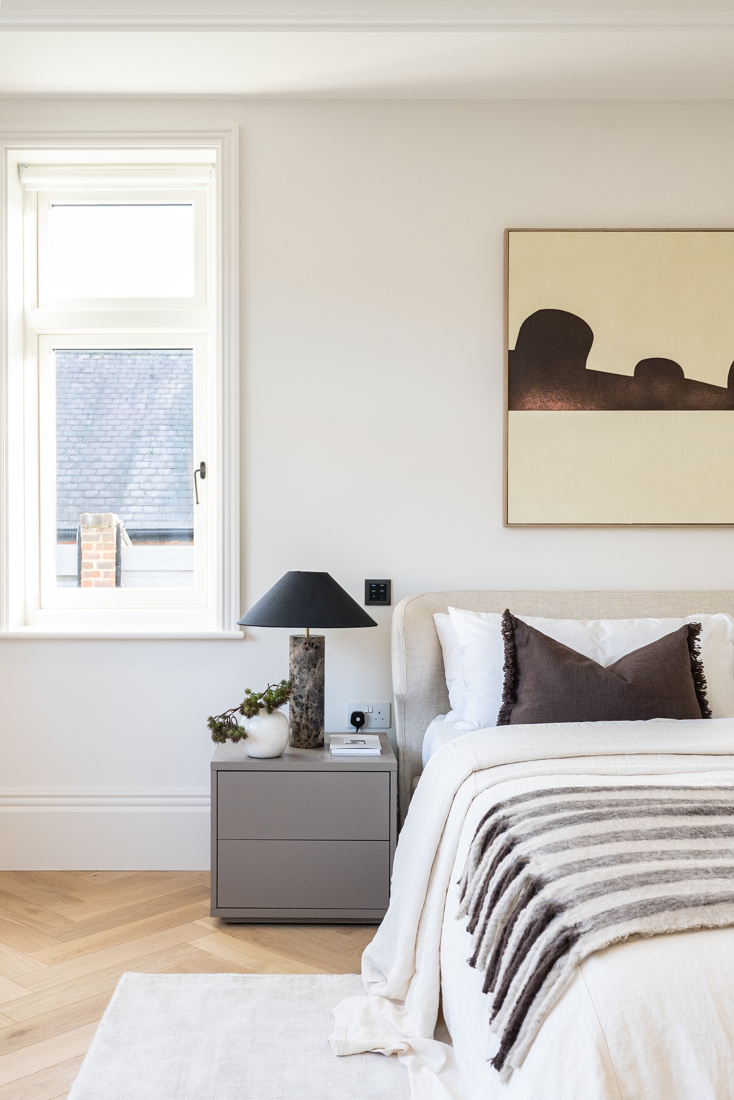 A bedroom with a window on the left, a gray nightstand with a black lamp, a white vase with greenery, and a book. The bed has white linens and a brown pillow, with a striped blanket. A painting hangs above the bed.