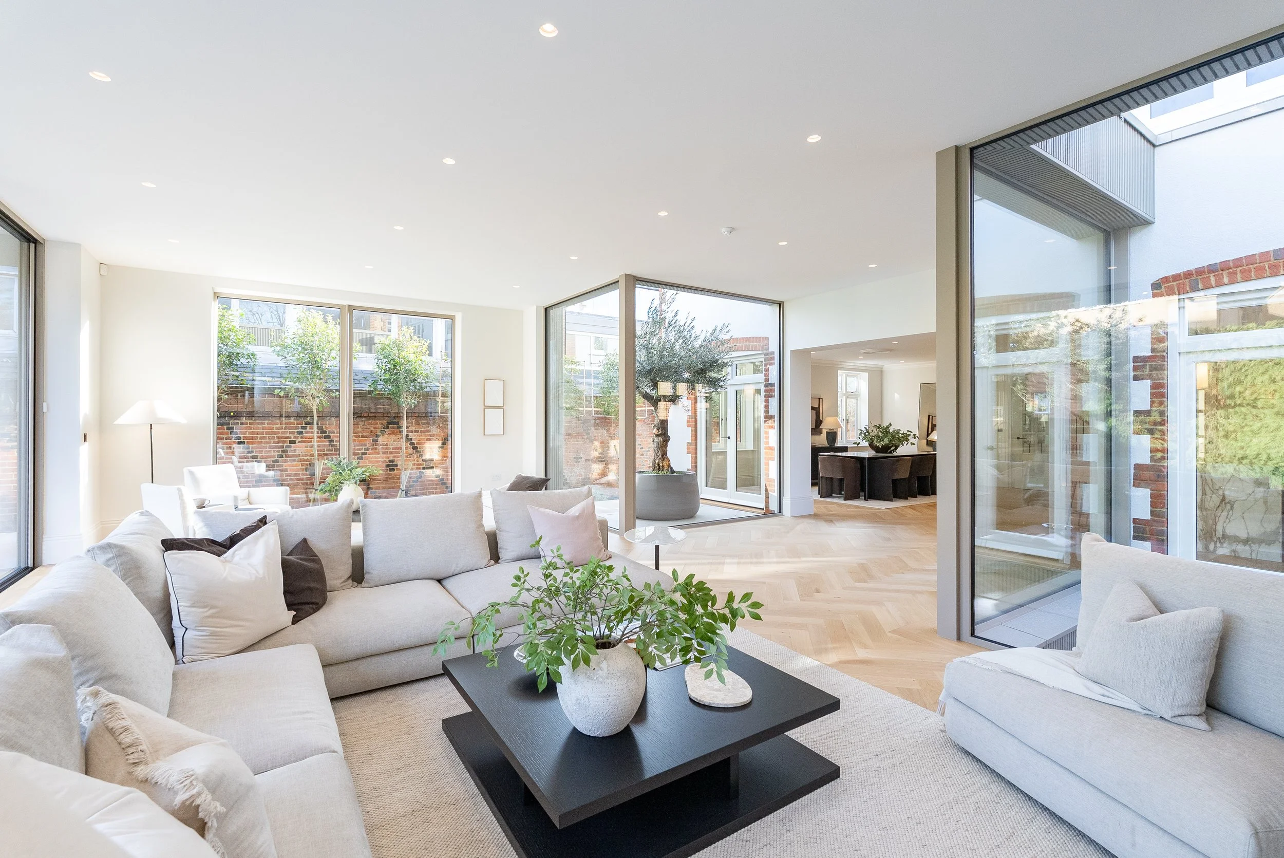 Bright, modern living room with large windows, white sofas, a black coffee table with a potted plant, and an open view to a patio with a tree.