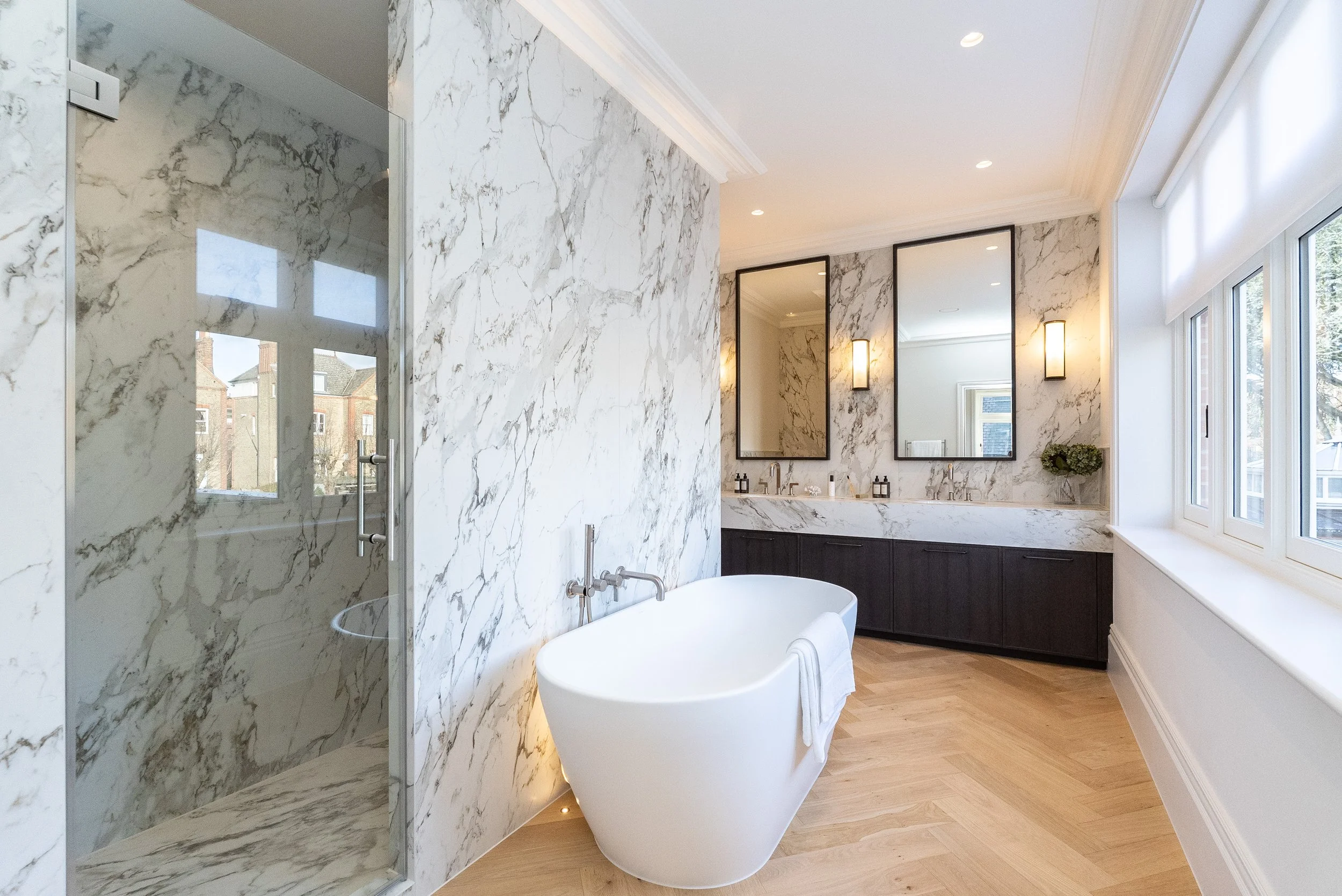 Modern bathroom with marble walls, a white freestanding bathtub, a glass-enclosed shower, and vanity mirrors above dual sinks. Large window with white blinds, wood flooring, and wall-mounted lights.