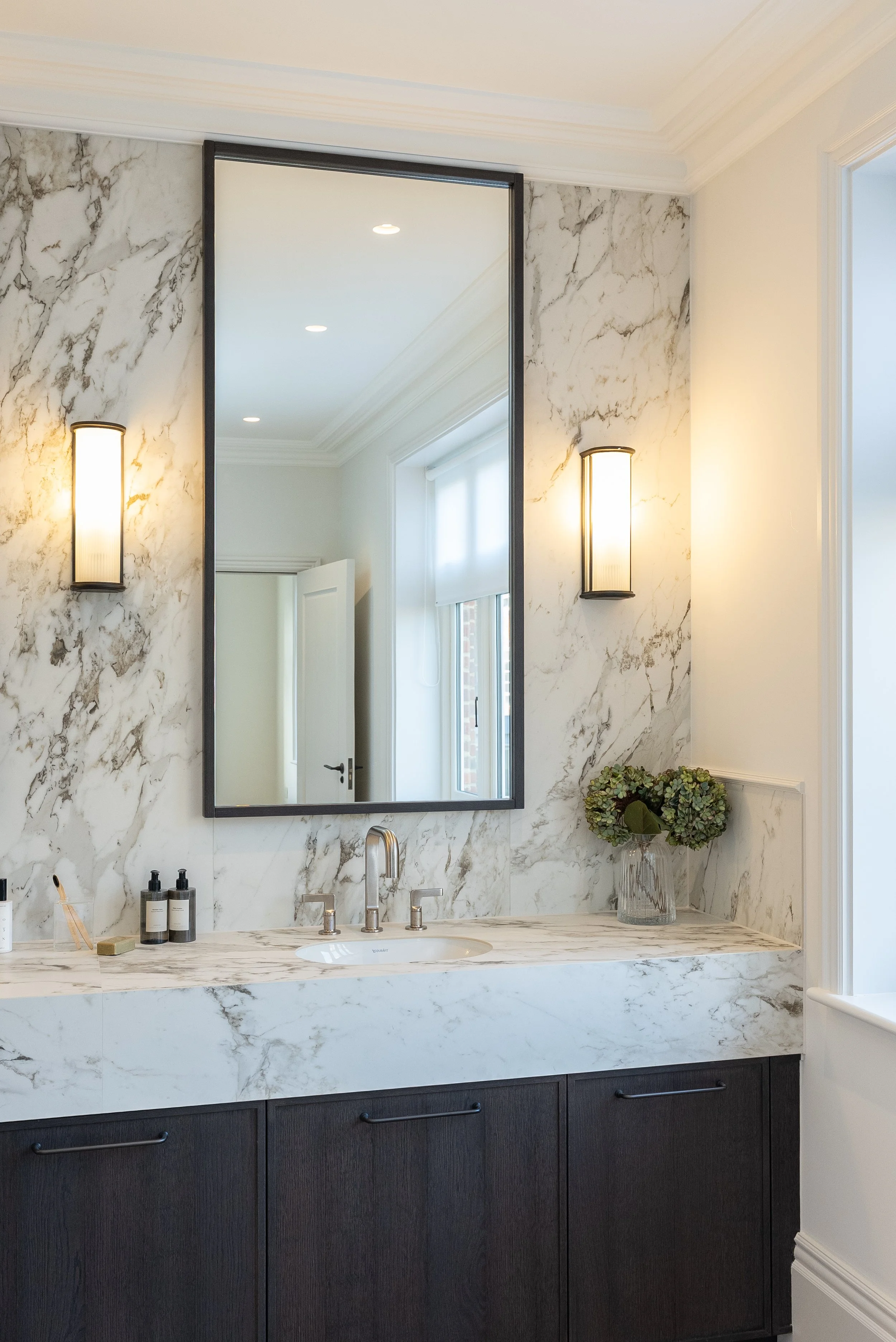 Bathroom vanity with marble countertop, dark wood cabinets, large mirror, wall-mounted lights, and a vase of green hydrangeas.
