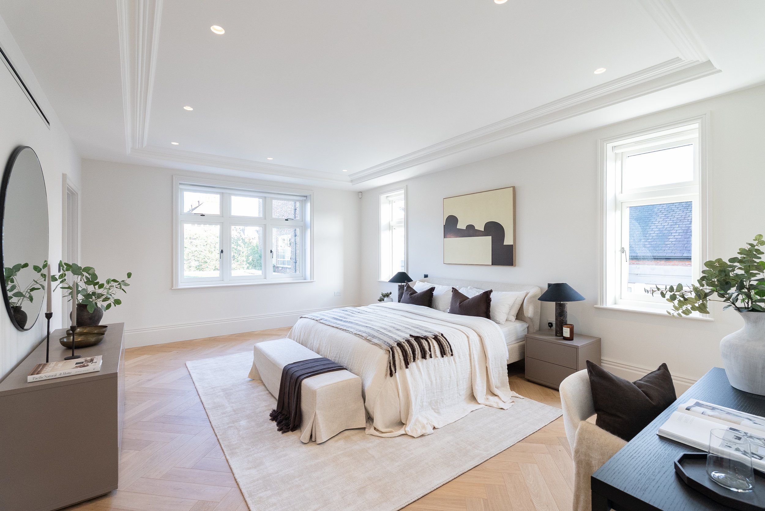 A bright, modern bedroom with white walls, large windows, wooden flooring, a bed with white and brown bedding, black bedside lamps, and a piece of abstract art on the wall.
