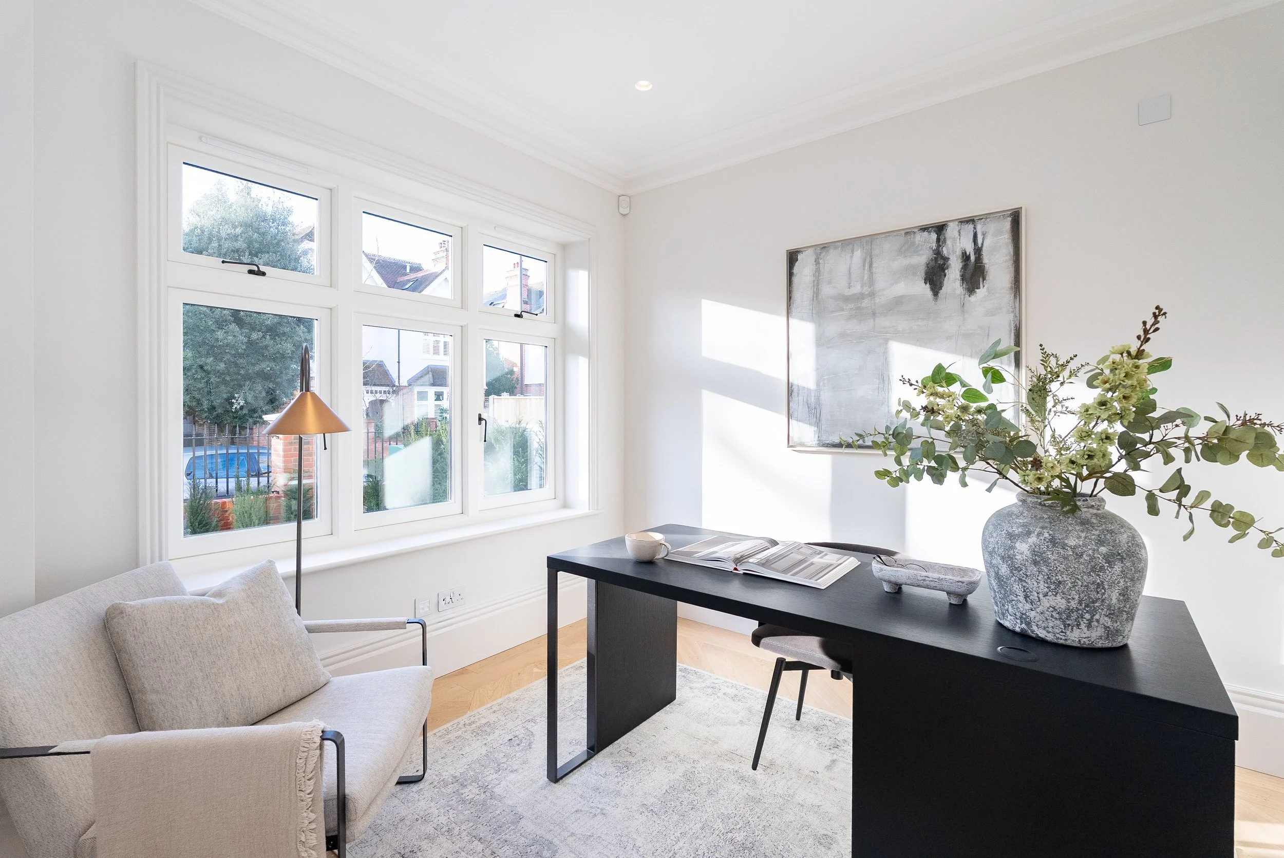 Bright modern home office with large window, black desk, vase with greenery, and abstract wall art on white walls.