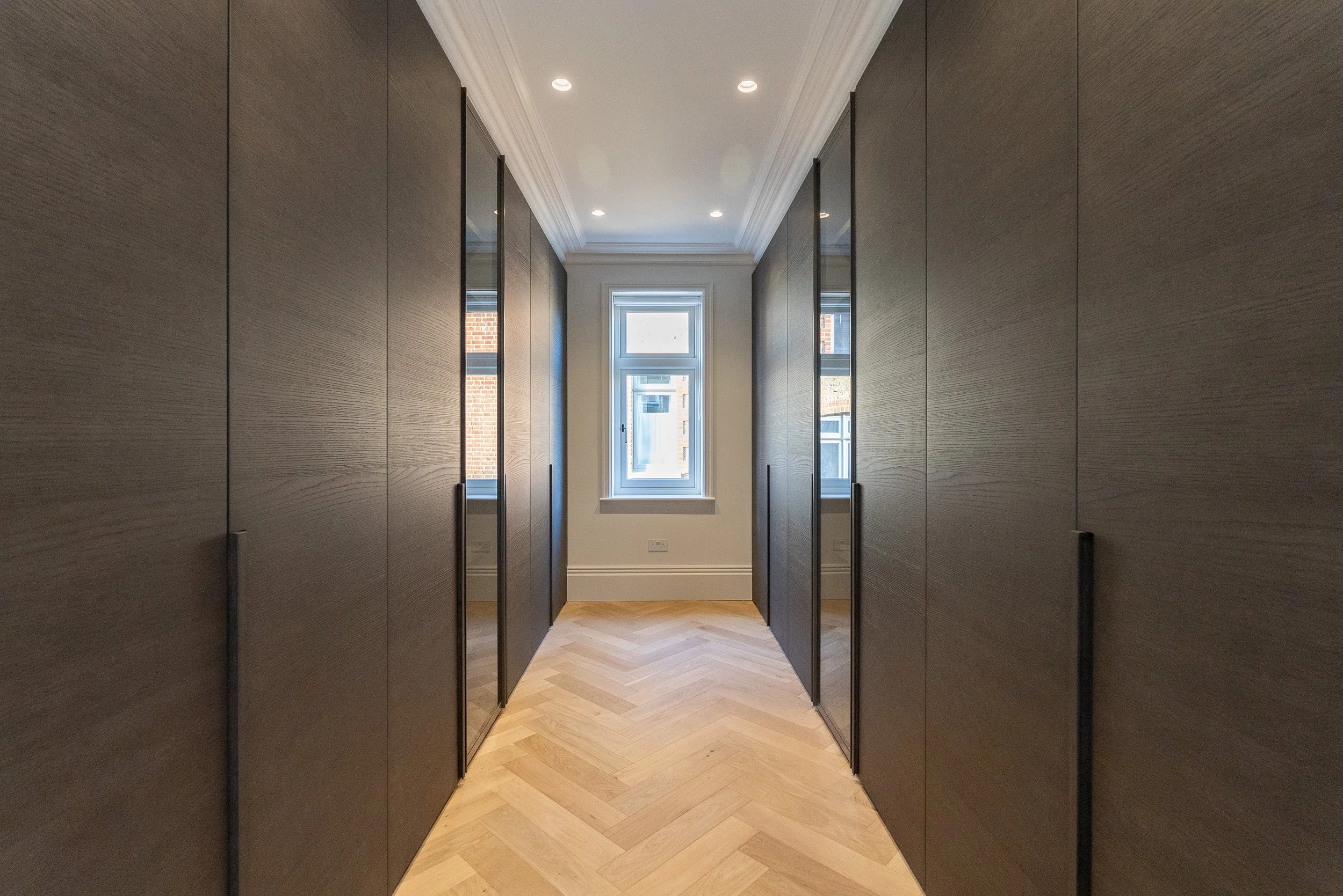 Interior hallway with dark wooden cabinets or closets on either side, a window at the end, and a light-colored herringbone floor.
