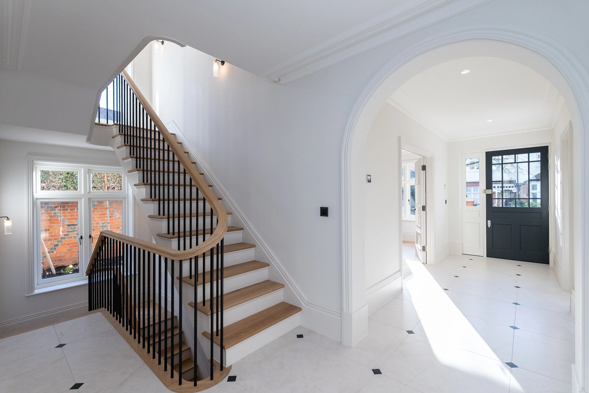 Interior of a modern house entryway with white walls, tiled floor with small diamond accents, a curved staircase with wooden steps and black metal railing, a black front door with glass panels, and a small window showing a brick wall outside.