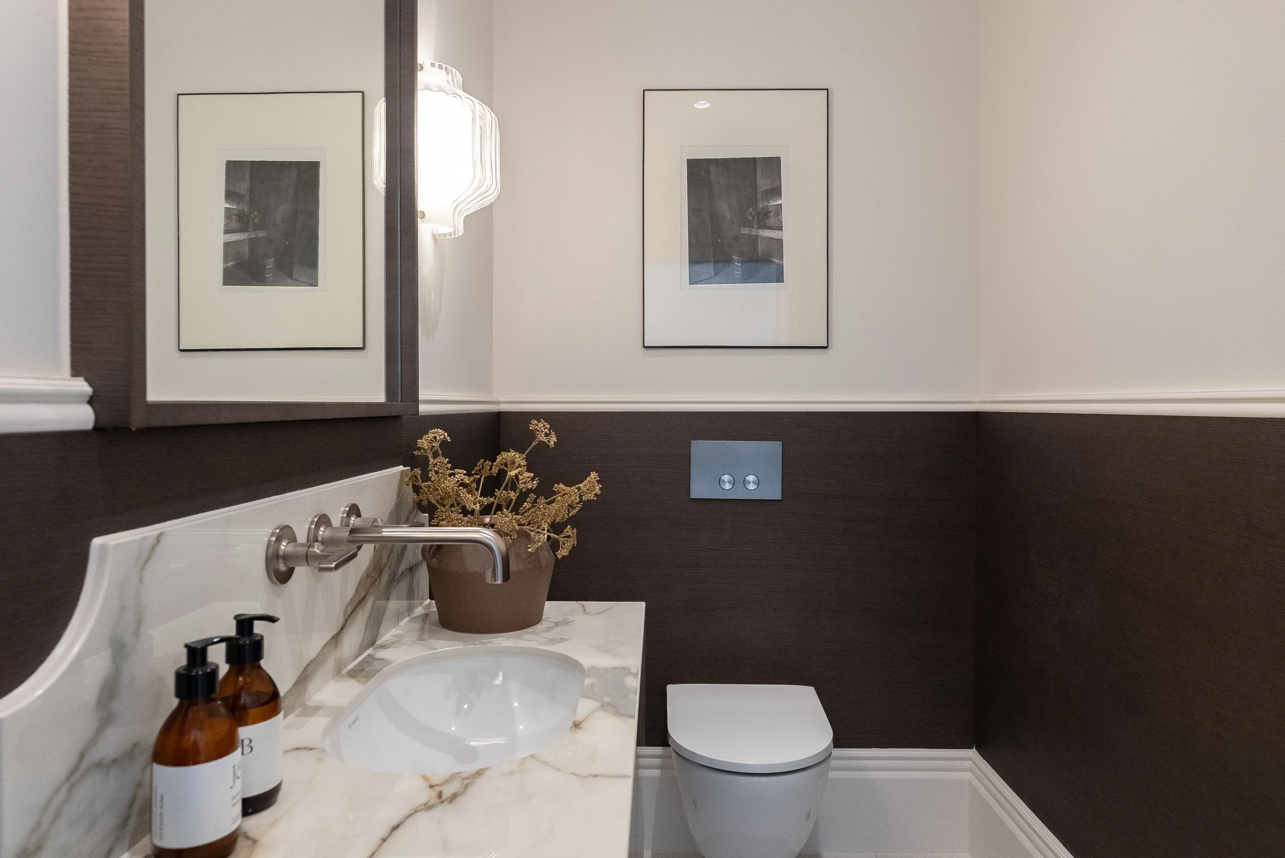 A modern bathroom with a marble countertop, a sink, two brown soap dispensers, a potted plant, a mirror, two framed photographs, a wall-mounted light fixture, and a toilet.