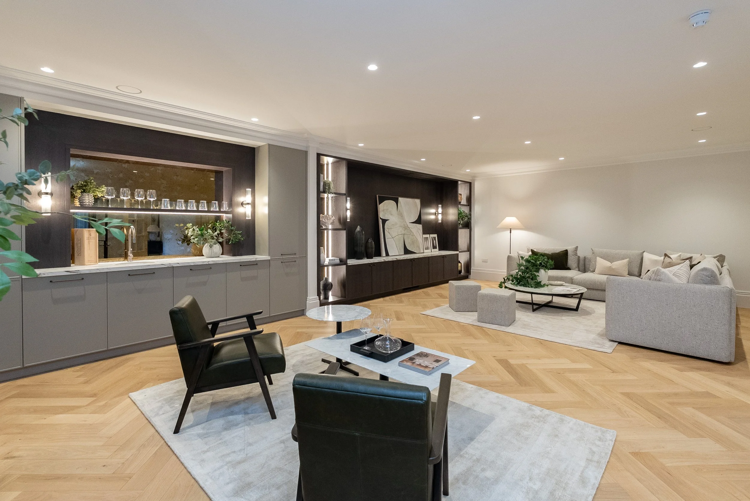 Modern living room with gray and black furniture, wooden floors, and minimalist decor, including a beige sectional sofa, black armchairs, a white marble coffee table, and a built-in bar area with glassware and plants.