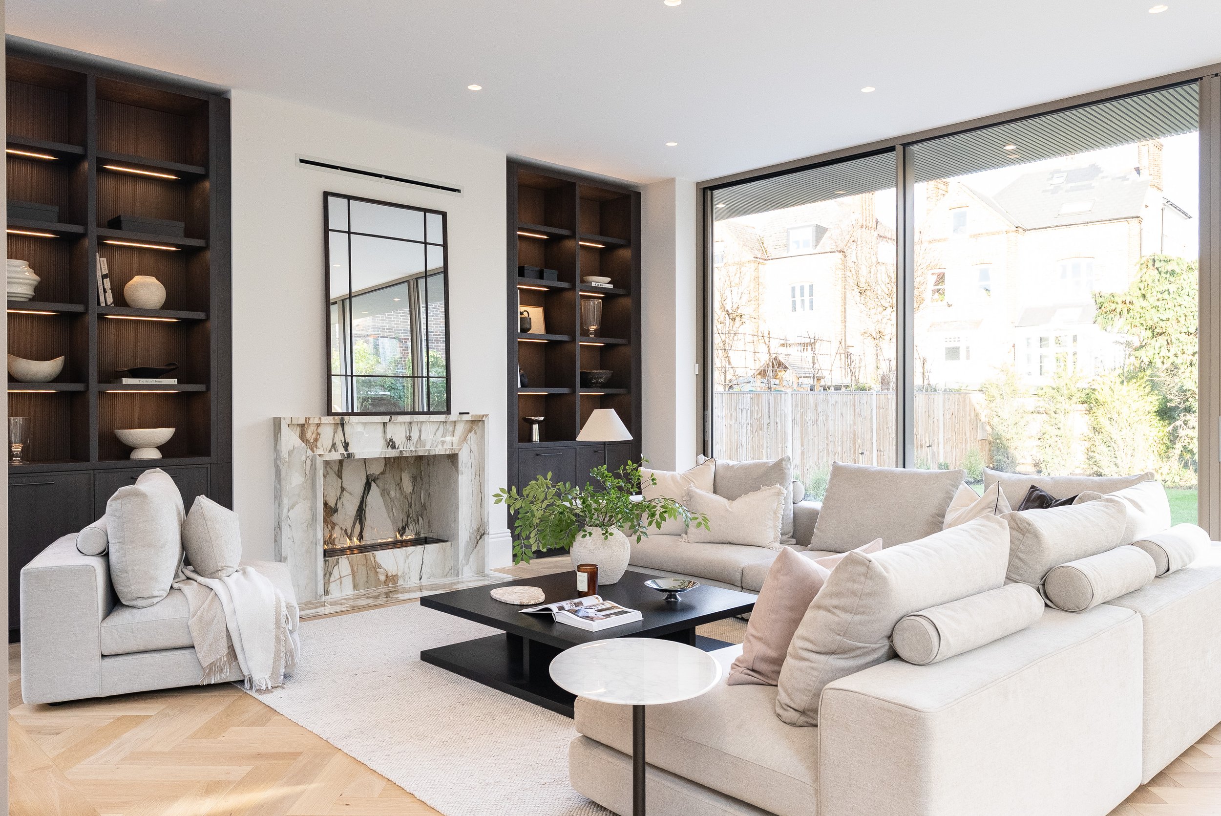Modern living room with large window, white sofas, black coffee table, marble fireplace, and built-in dark wood shelves.