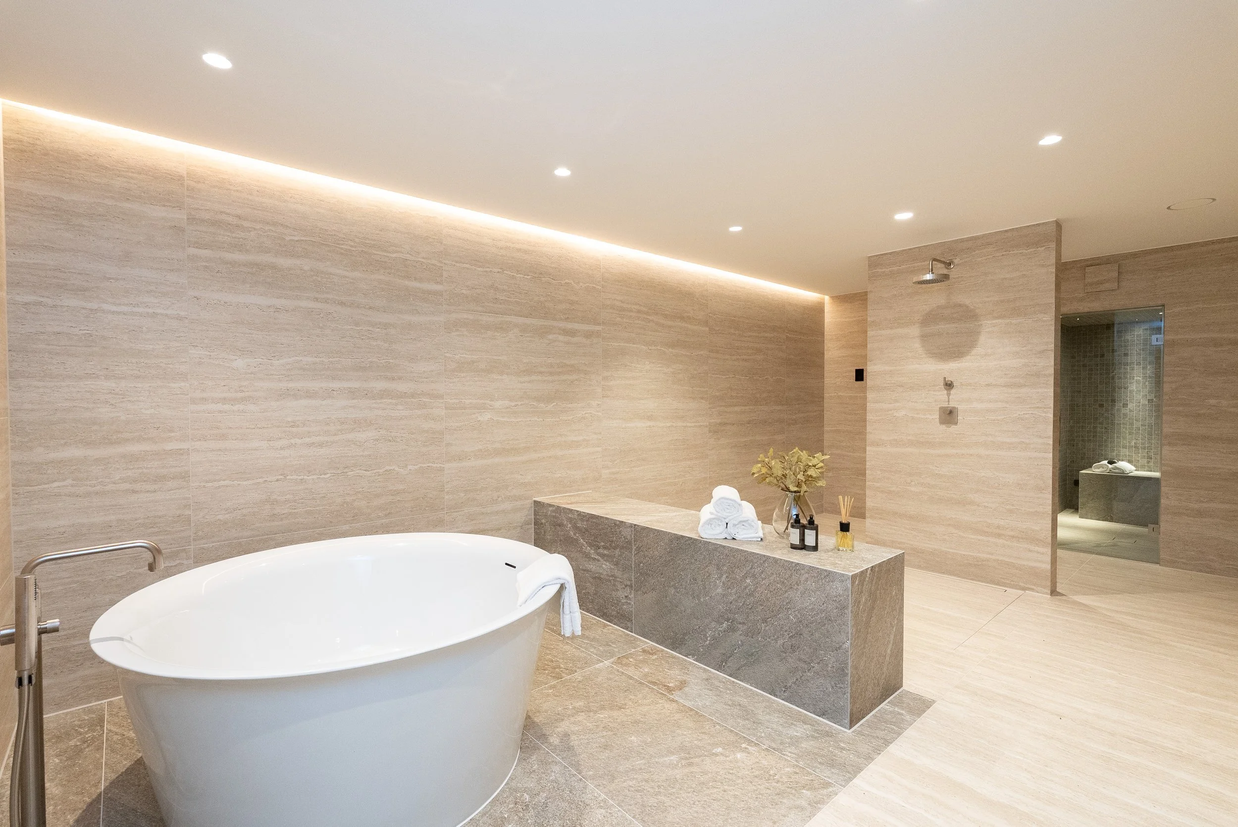 Modern bathroom with a freestanding white bathtub, beige tiled walls and floor, a small gray bench with towels and toiletries, and a separate shower area visible through an open doorway.