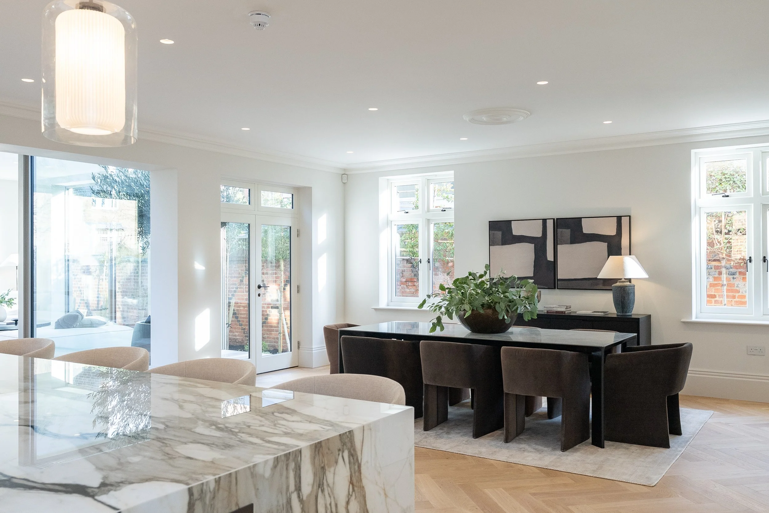 A modern, well-lit living and dining area with large windows and a door leading outside. The room has a black dining table with matching chairs, a black sideboard with a lamp and artwork, and a marble kitchen island in the foreground.
