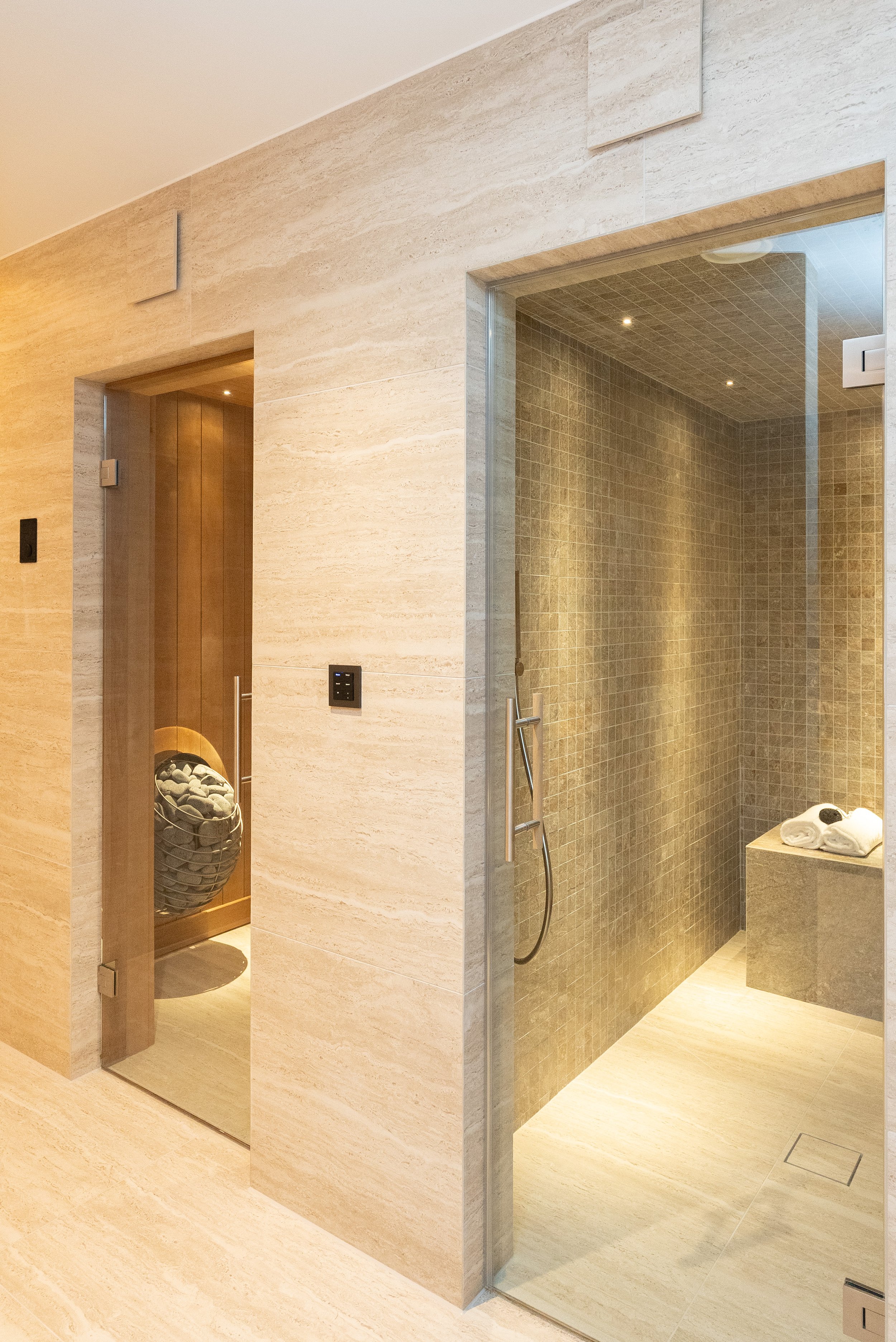 Modern bathroom with a glass shower enclosure, beige tiles, and a wooden sauna room.