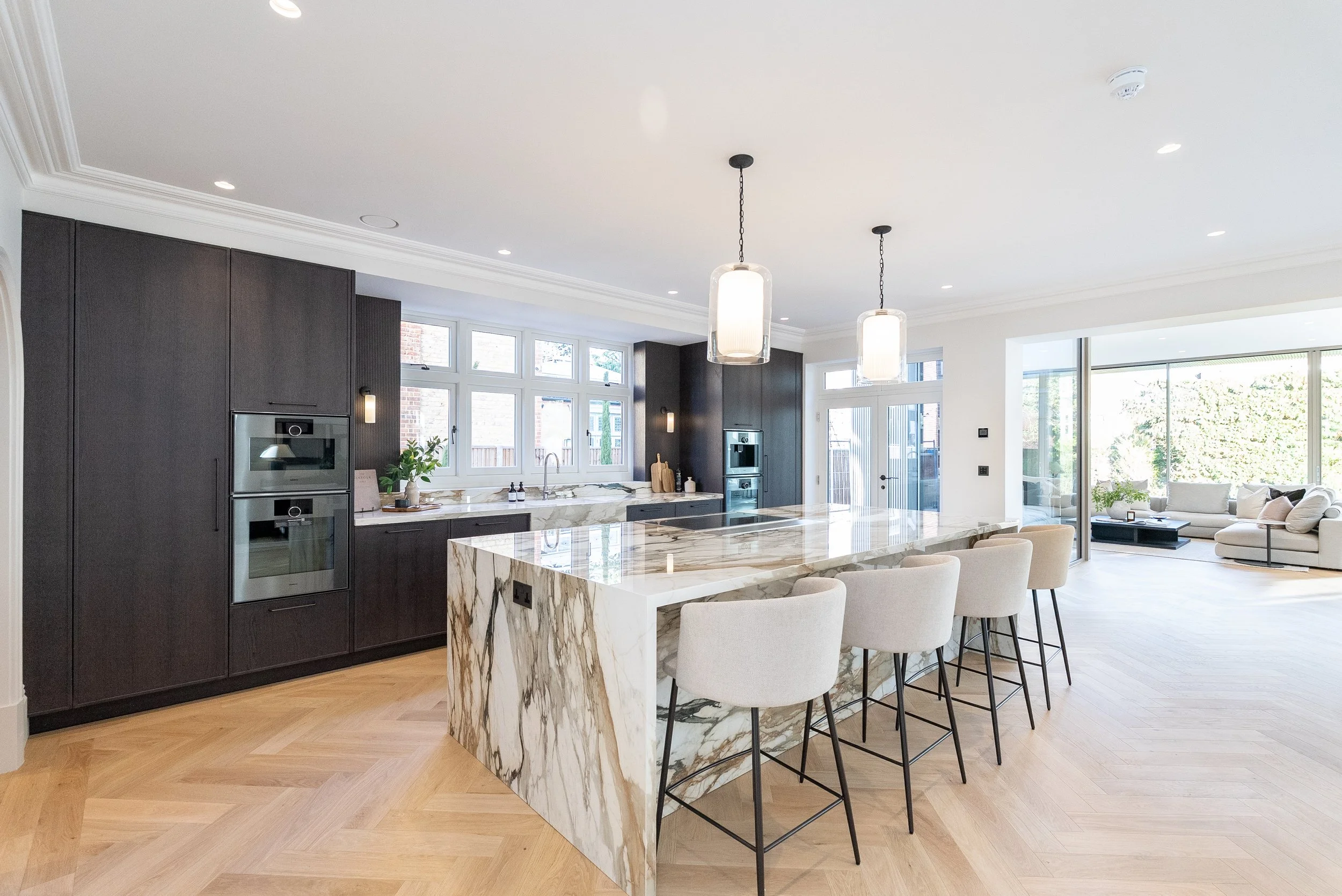 Modern open-concept kitchen with dark cabinetry, a marble island, and a living space with large windows and a white sofa.