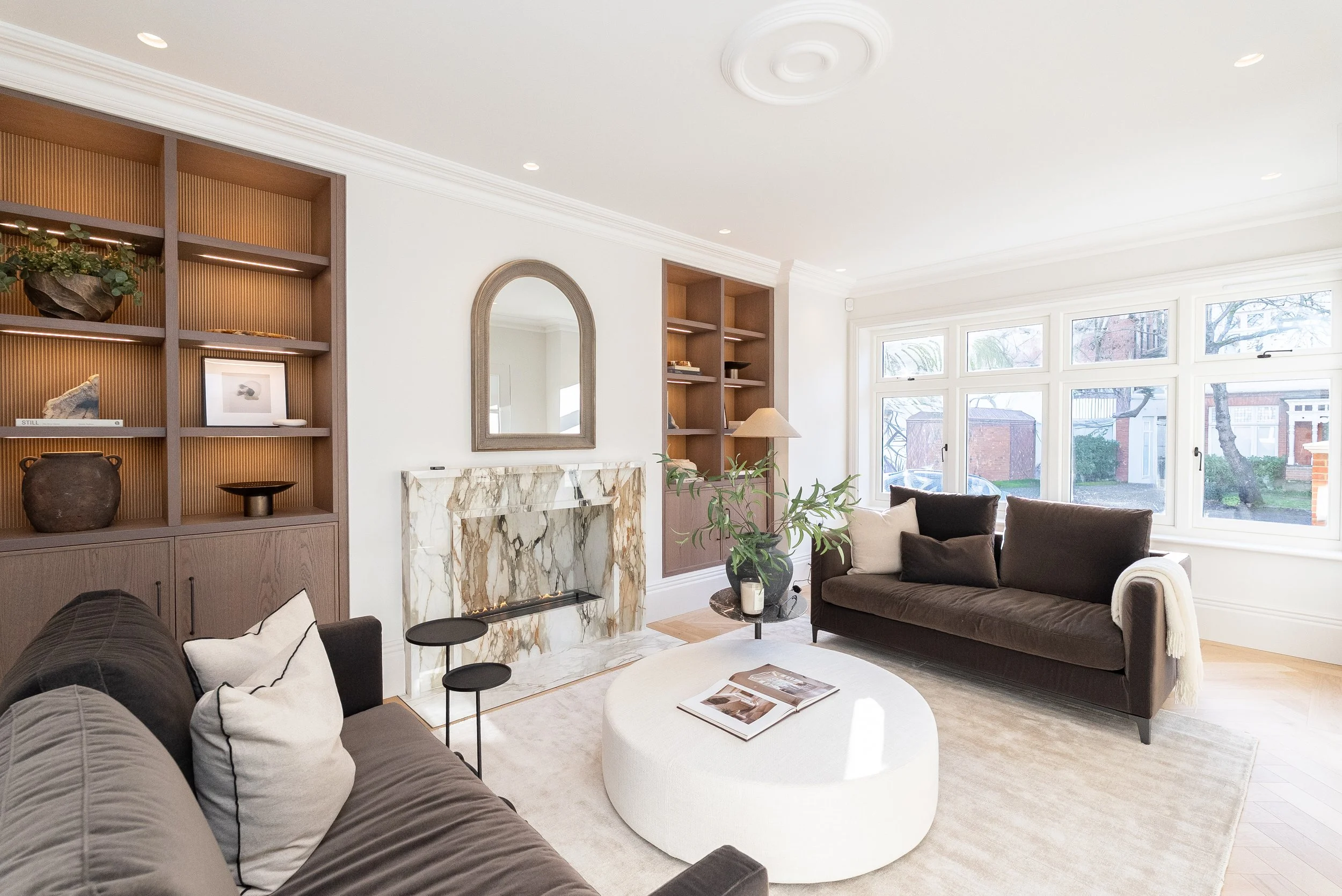 Bright living room with large bay window, brown and white sofas, marble fireplace, built-in wooden shelves with decor, potted plant, round white coffee table, and light-colored rug.