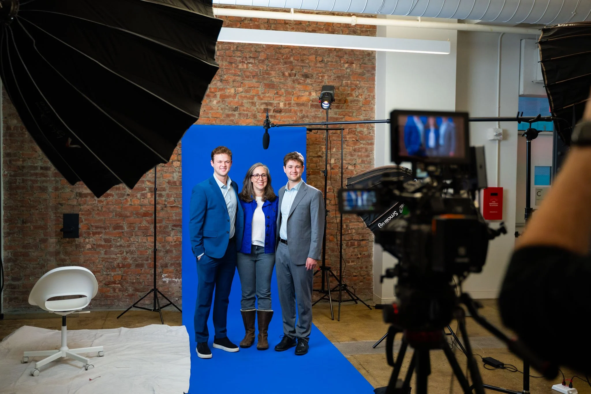 A woman and two men stand in front of a blue screen in a studio with professional lighting and equipment, smiling at the camera.