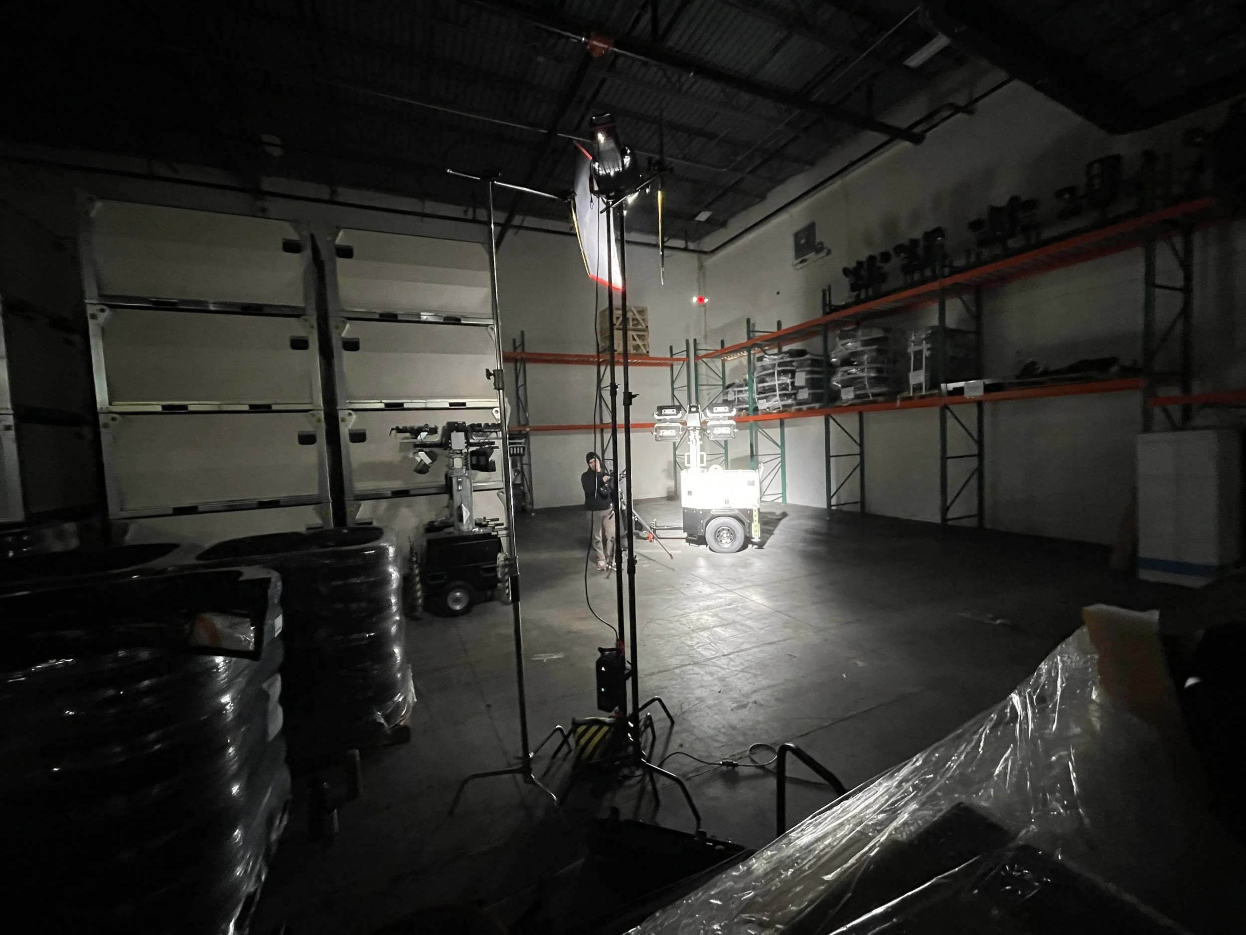 A photography studio with lighting equipment, a person working near a portable light source, and shelves with various items in the background.