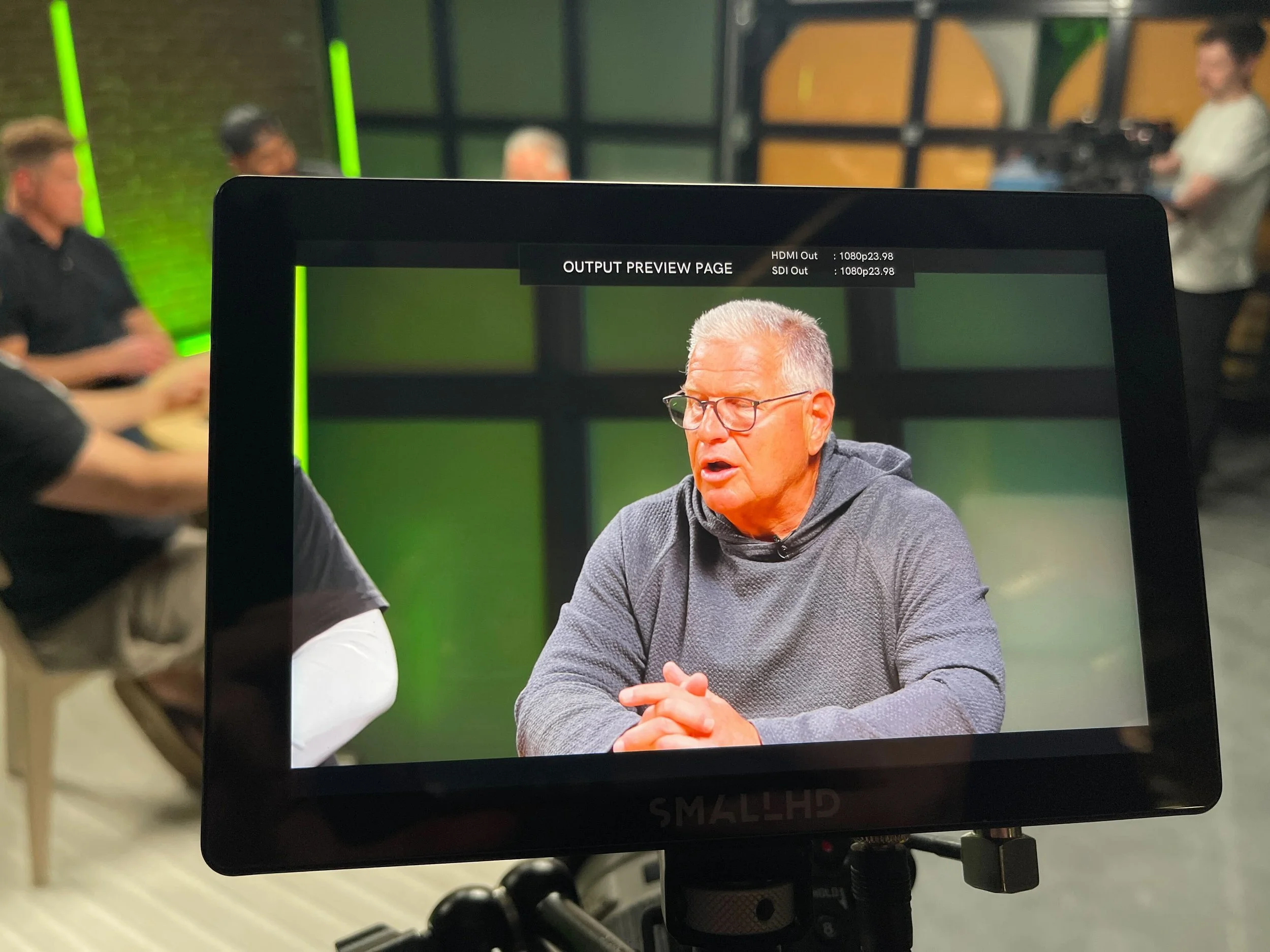 A professional video camera monitor displaying a scene with an older man with glasses in a gray hoodie, talking and gesturing with his hands, while three other people are blurred in the background.