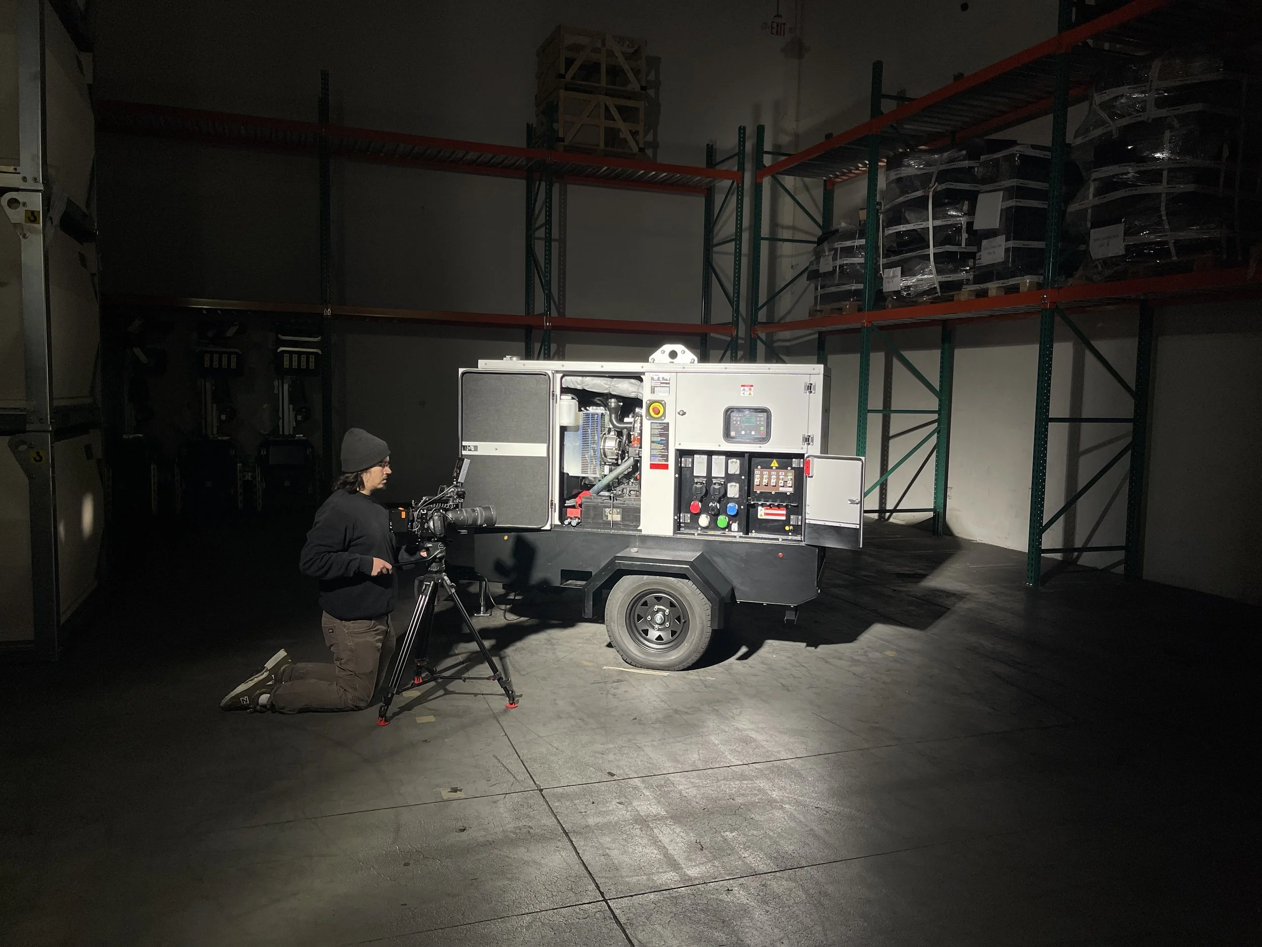 A person kneeling on the floor operating a camera mounted on a tripod, filming a portable generator in an industrial warehouse with shelves in the background.