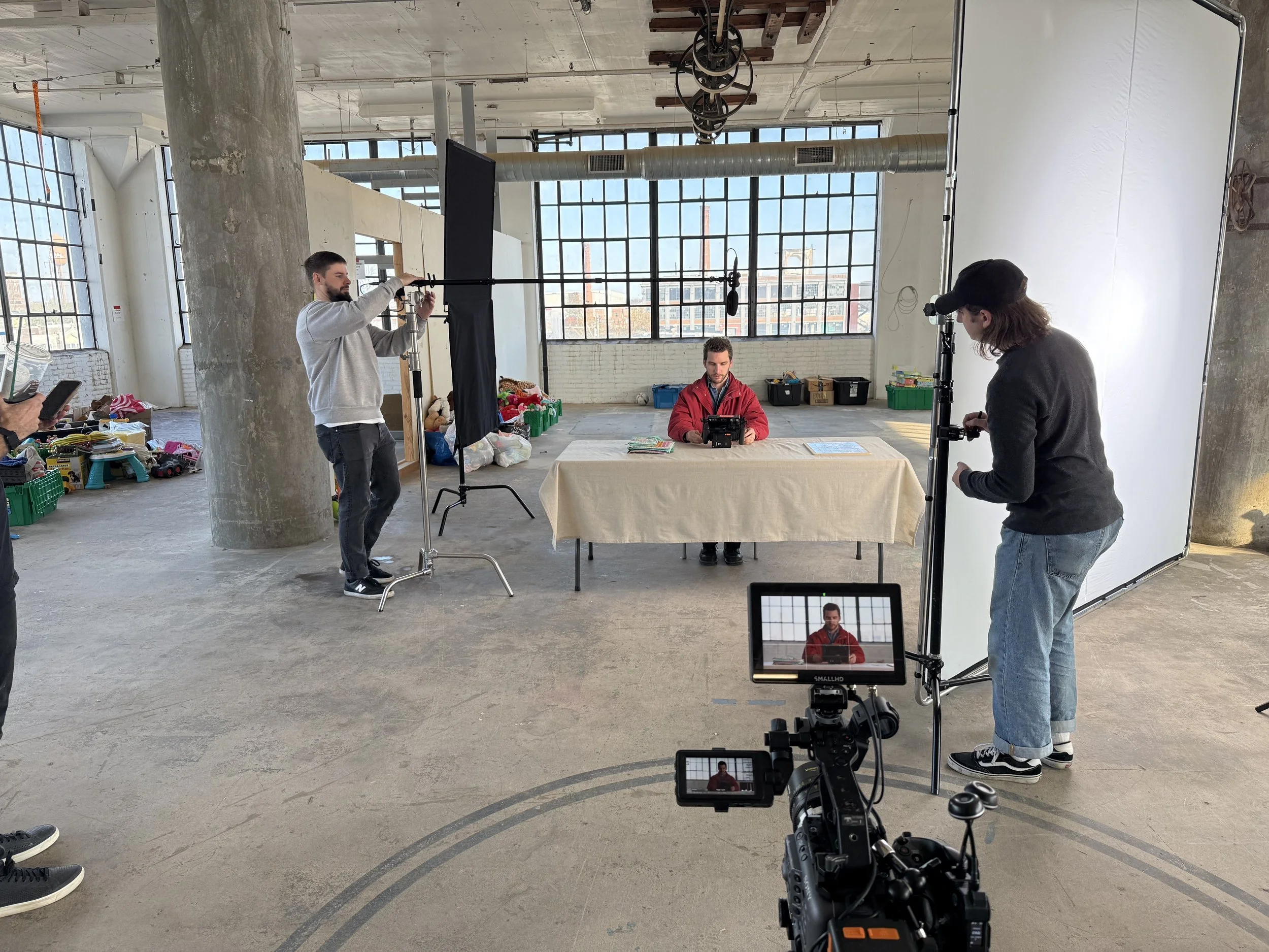 A man in a red jacket video blogs at a table with a beige tablecloth, while a camera crew films him in an industrial-style studio with large windows, a concrete pillar, and a white backdrop.