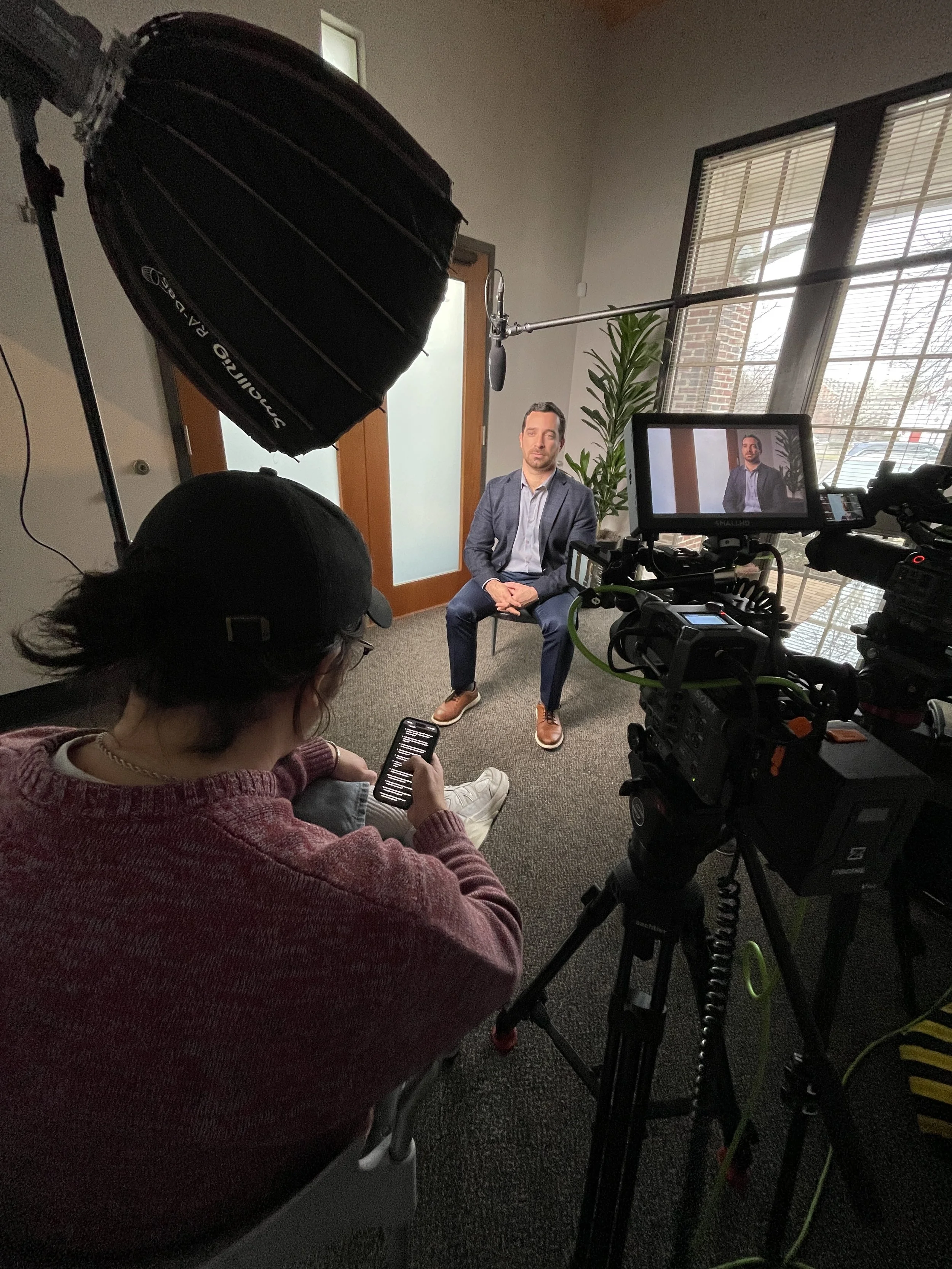 A man in a gray blazer and white shirt being filmed in a studio with professional lighting and camera equipment, sitting on a chair in front of a large window and potted plant, with a crew member using a smartphone nearby.