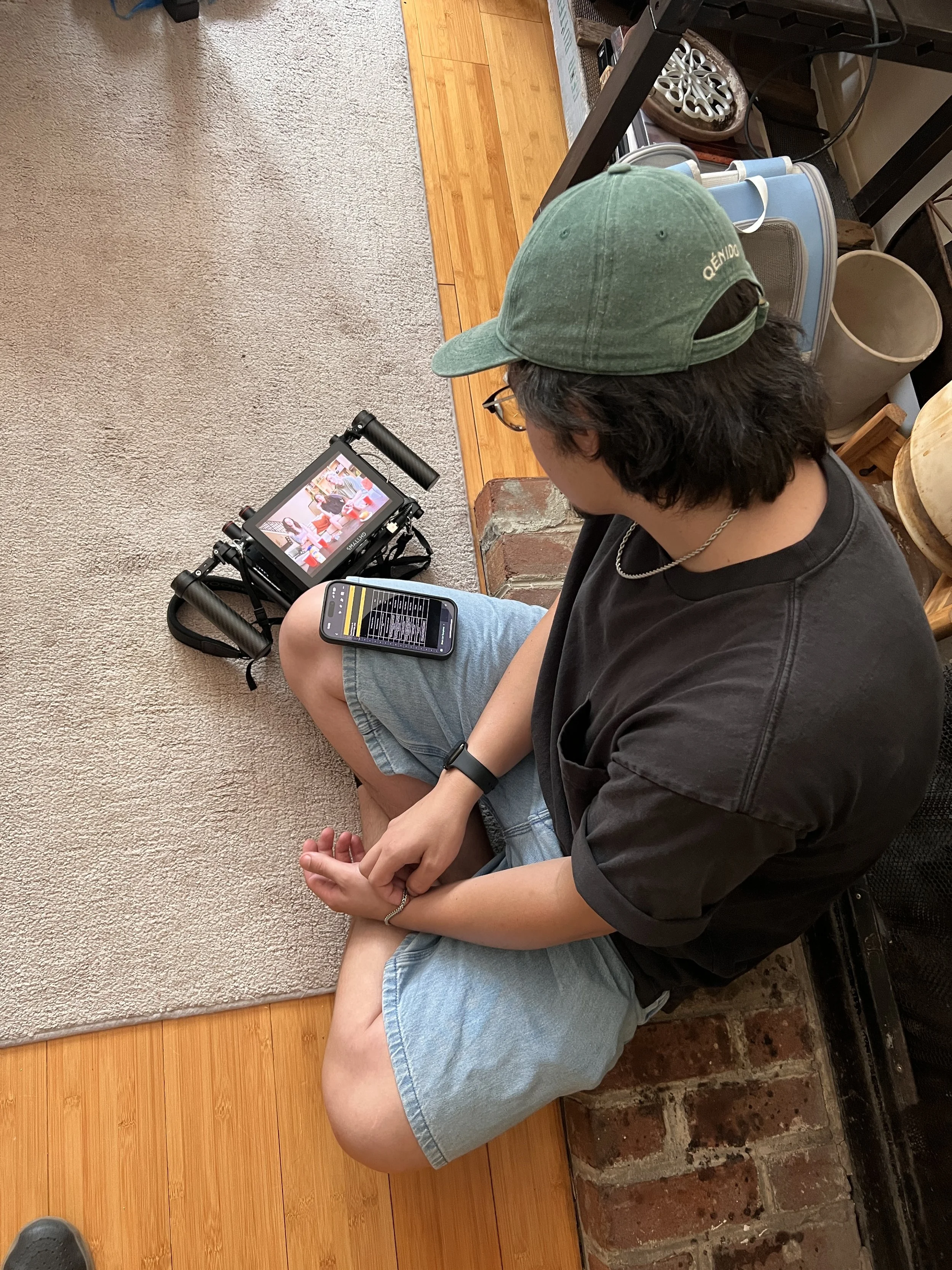 A person sitting cross-legged on the floor, looking at a small camera and a smartphone, with a TV screen showing a scene of several people in the background.