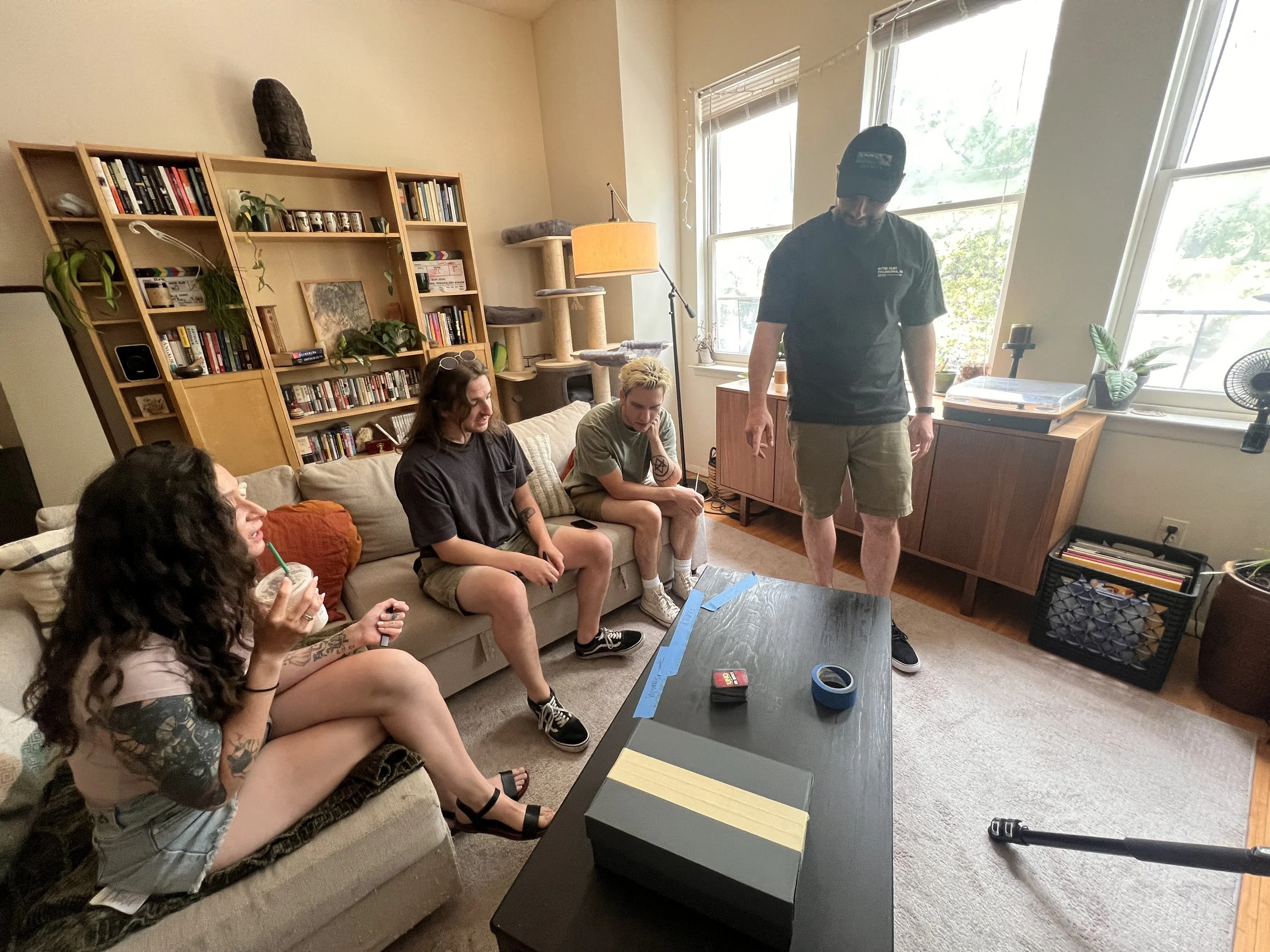 A group of four young women and one man in a living room. The women sit on a beige couch, one drinking a Starbucks frappuccino, another with glasses and tattooed arms, and two with short hair. The man stands near a window, looking down at a black table with game pieces and tape. The room has a bookshelf, cat tree, and plants near the windows.