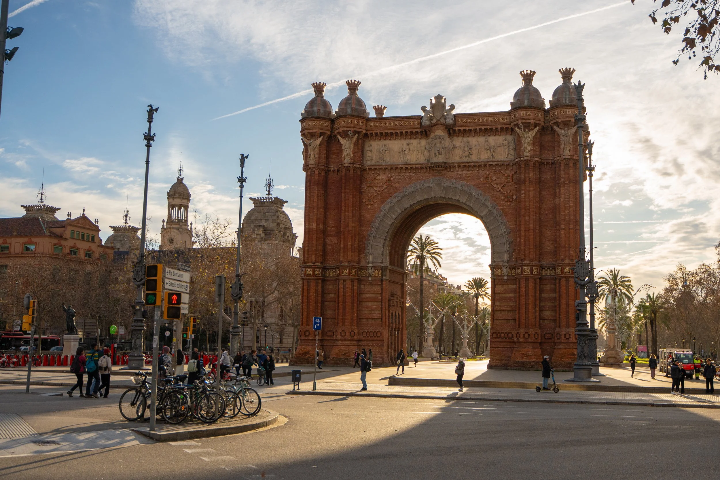 The entrance arch of the Ciudadela park in Barcelona, Spain, with people walking and biking nearby, and palm trees and historic buildings in the background under a partly cloudy sky.