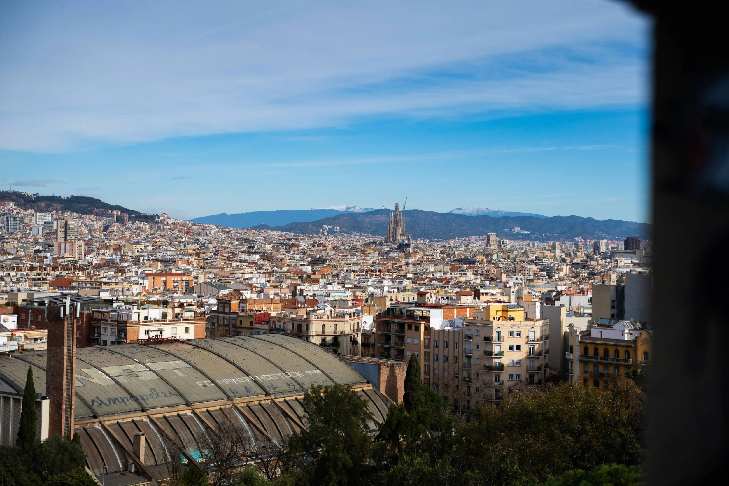 Cityscape of Barcelona with the Sagrada Familia basilica in the distance under a blue sky.