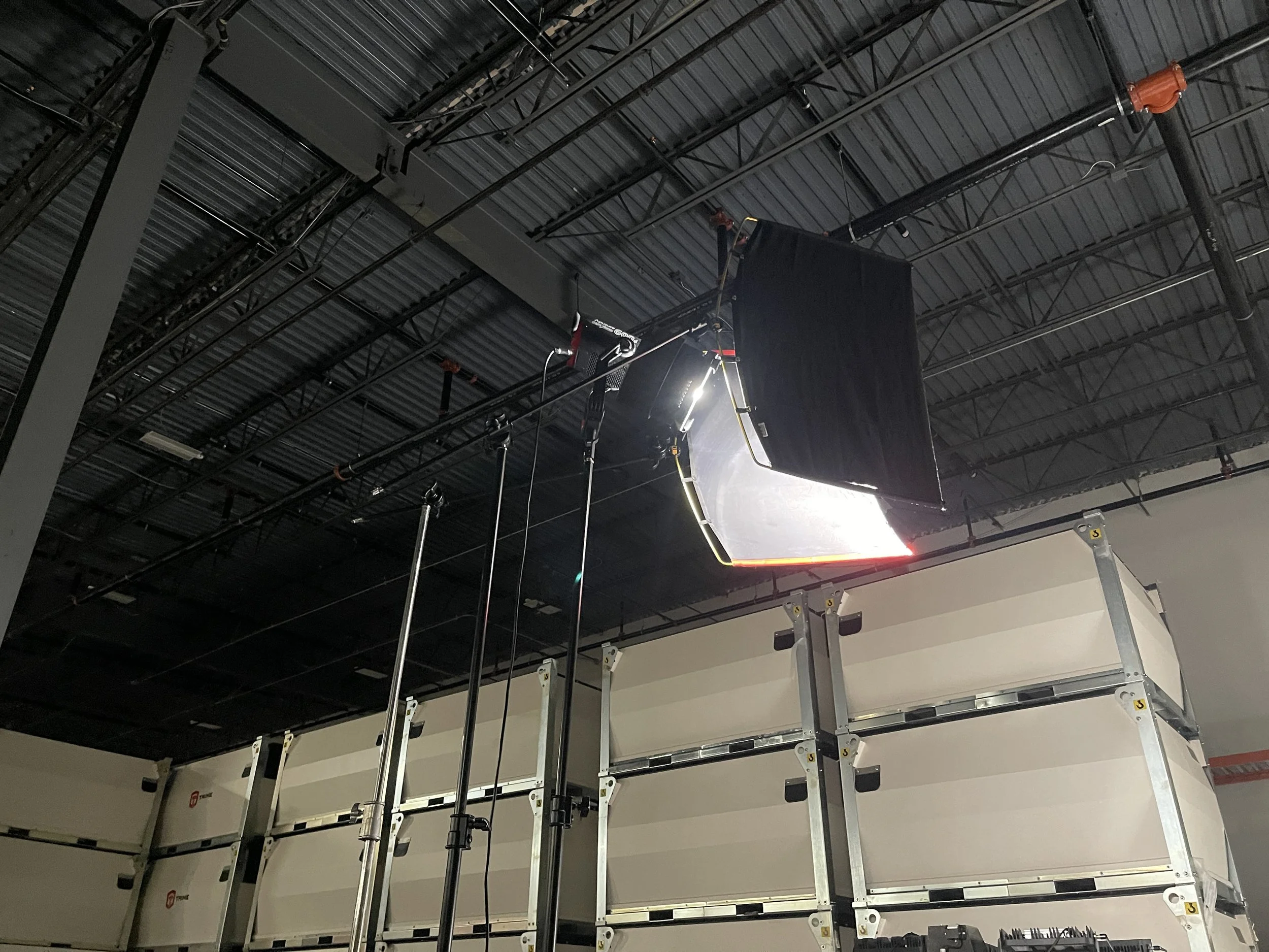 Photo of a professional studio lighting setup, including a large softbox and light stand, in a warehouse or studio with stacked storage containers.